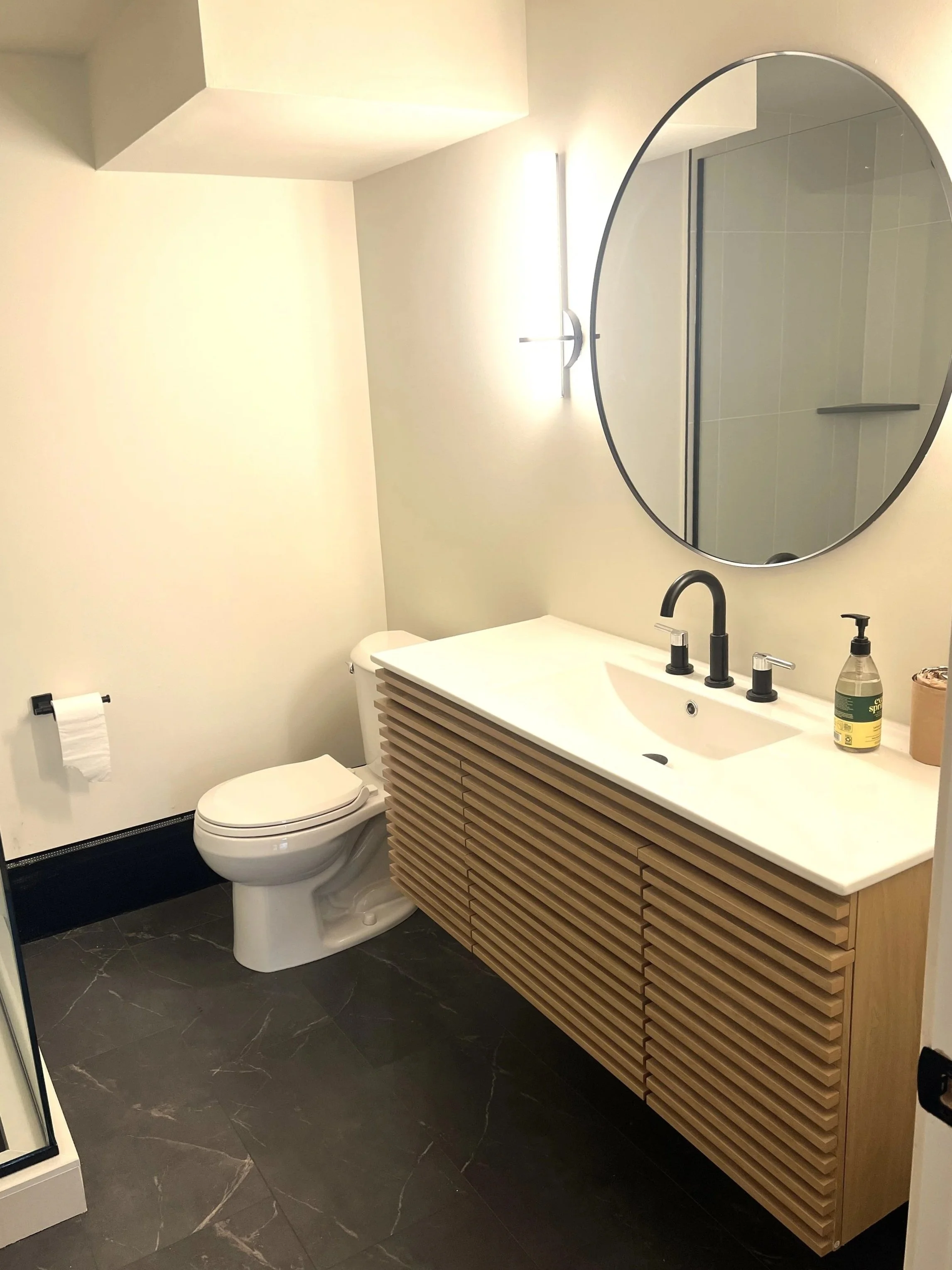 A modern bathroom with a white toilet, a wooden vanity with a white countertop and black fixtures, a large round mirror, a wall-mounted light, a green soap dispenser, and a towel holder with a white towel.