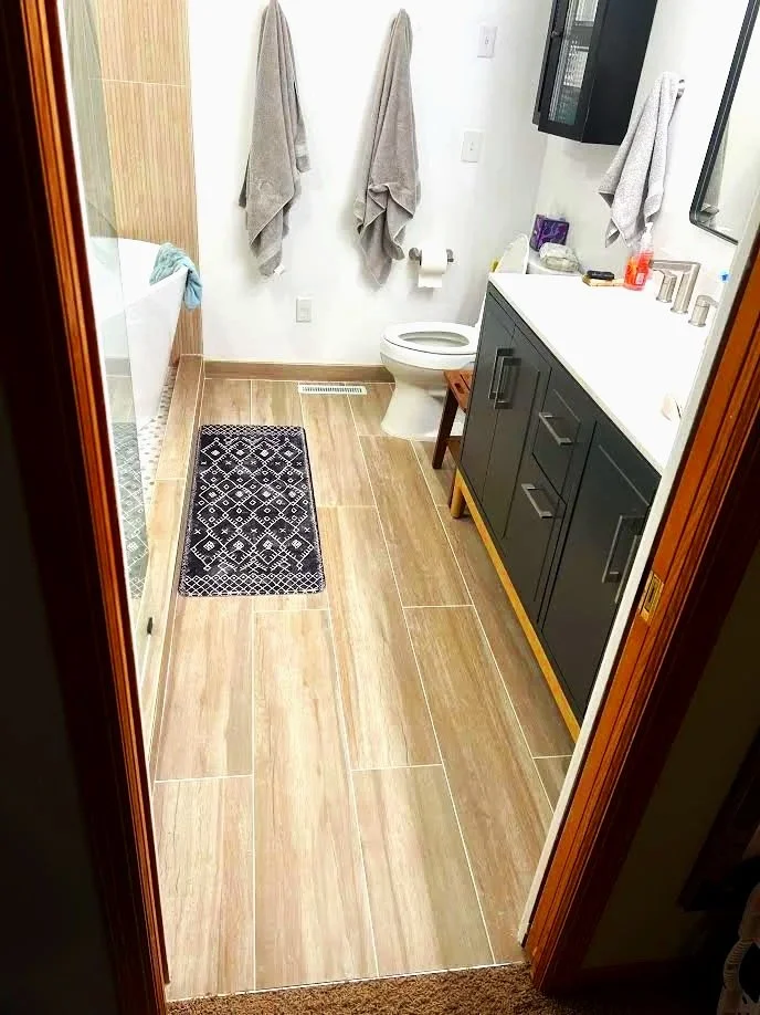 A bathroom with a wooden frame door, tan tile flooring, two gray towels hanging on the wall, a toilet, a black bathroom vanity with a white countertop, and a black and white patterned rug.