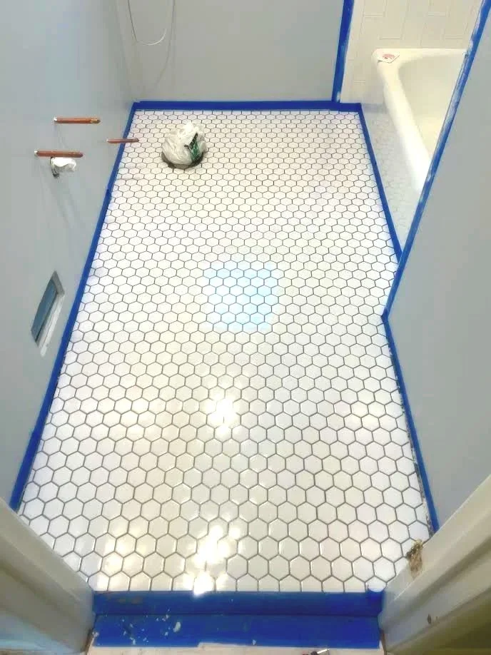 Bathroom with white hexagonal tile floor and blue painter's tape along the edges. A plastic bag is on the floor near the back wall, with a hole in the wall on the left side, and a partially visible bathtub on the right.