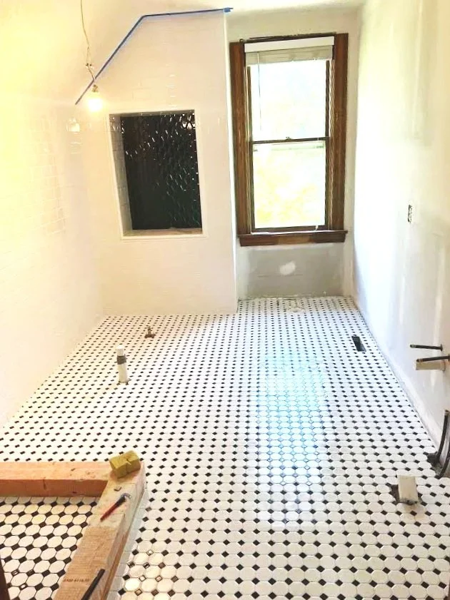Room under construction with black and white hexagon tile flooring, a window on the back wall, and visible plumbing pipes.