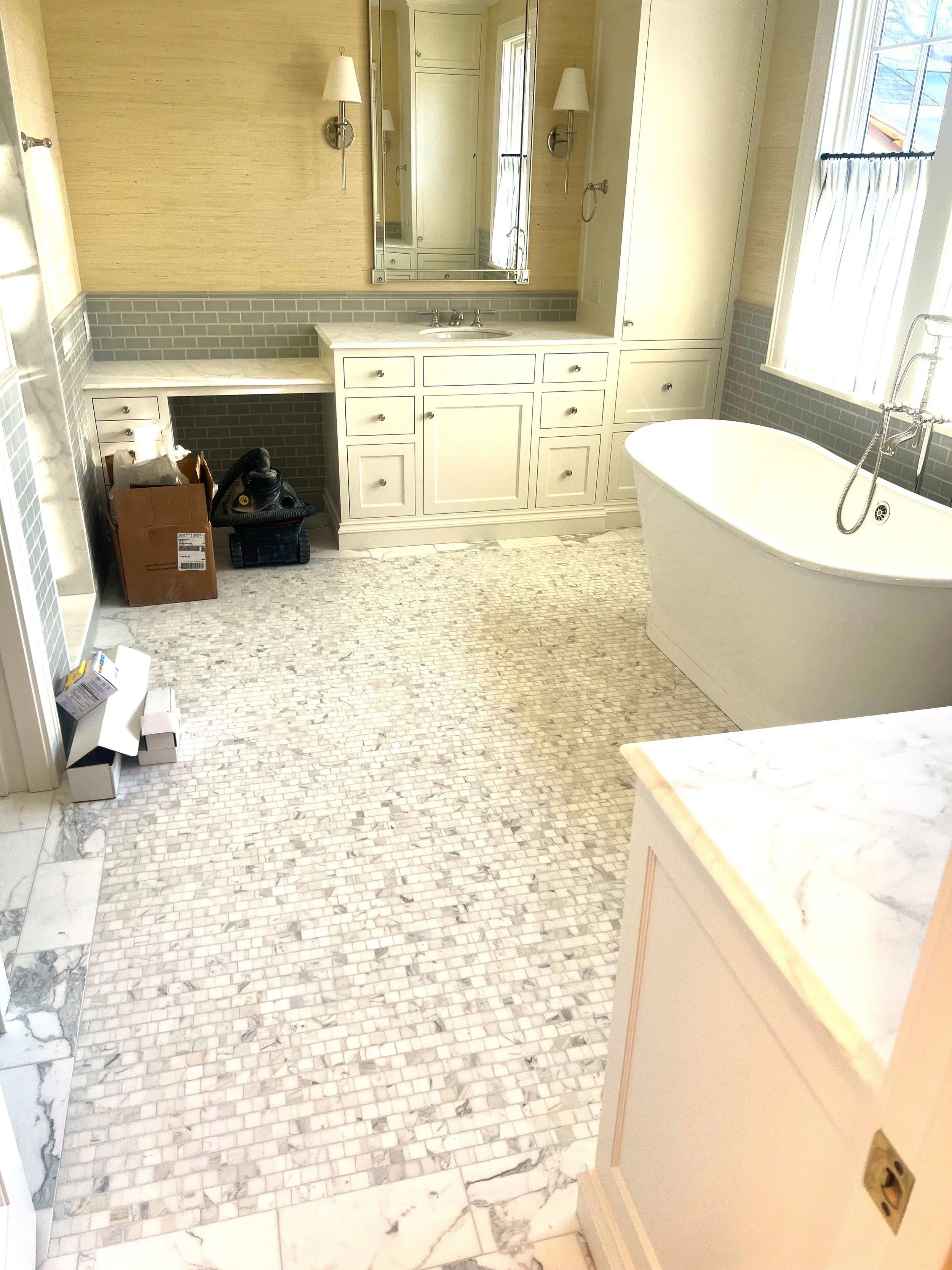 Bathroom with a freestanding bathtub, large window with curtains, vanity with mirror, and light fixtures, with floor tiles in a mosaic pattern.