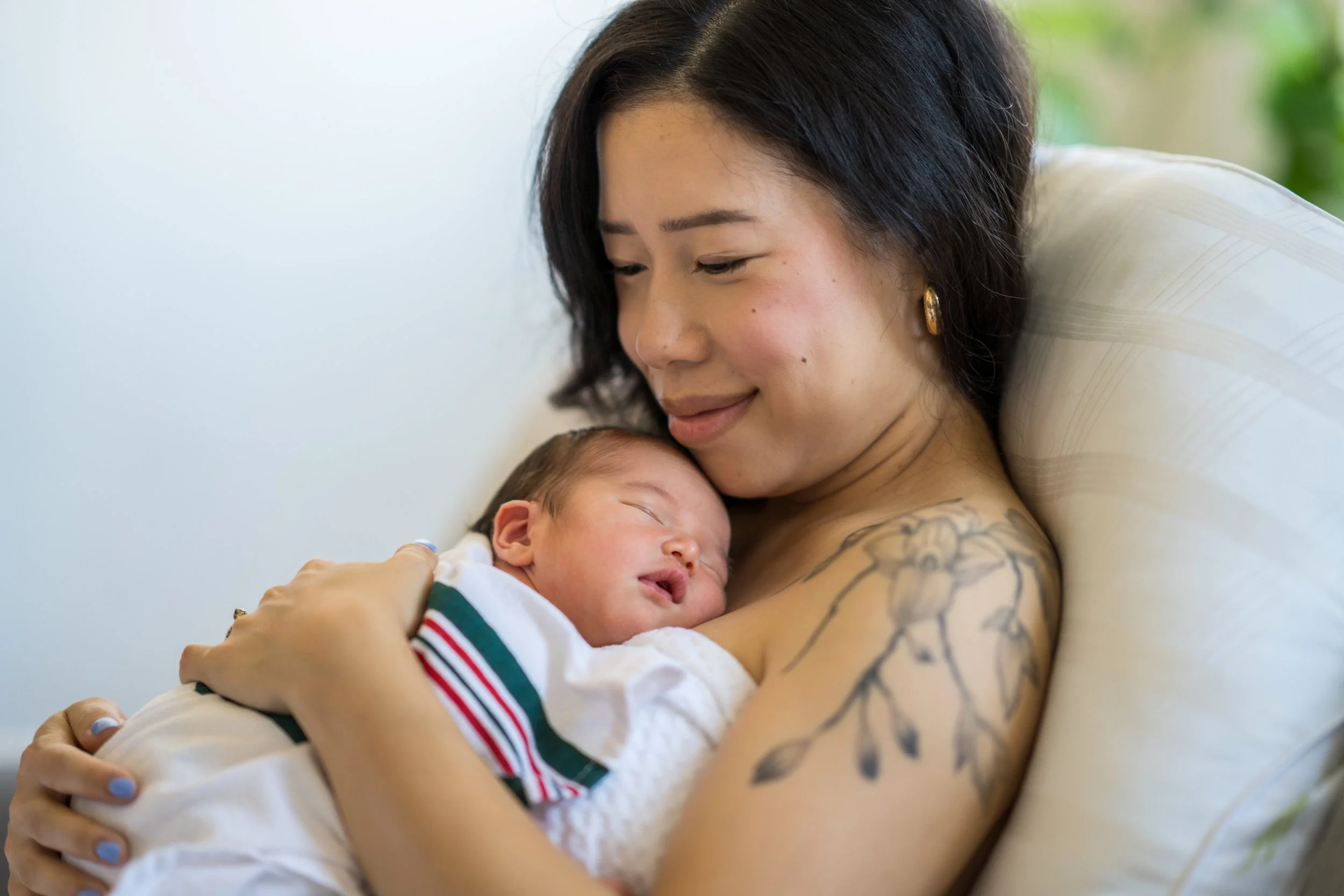 A woman with dark hair and tattoos on her arm gently holding a sleeping newborn baby, smiling softly while resting on a pillow.