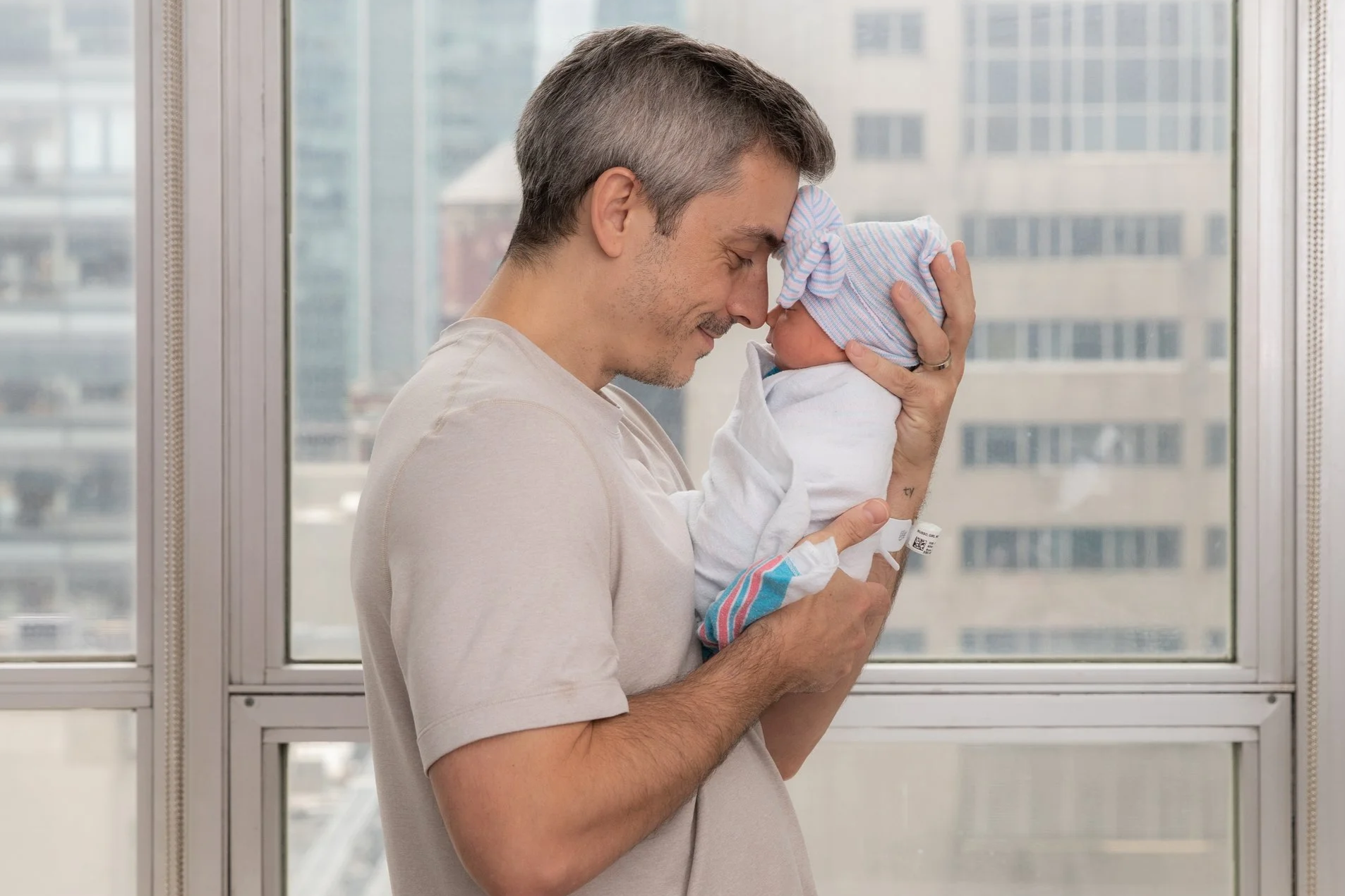 Man holding newborn baby with a hospital wristband, near a window in a high-rise building.