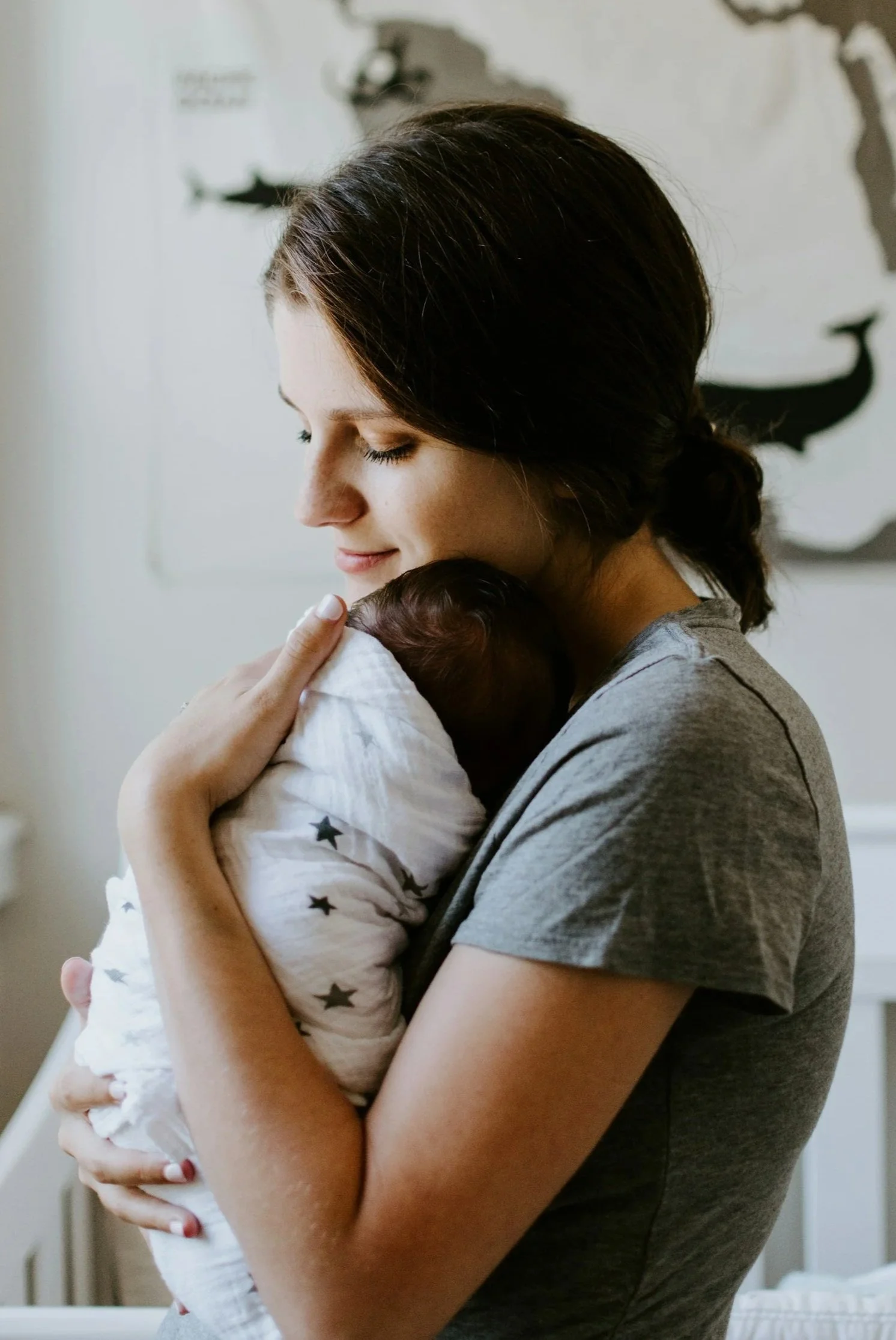A woman gently holding a sleeping newborn baby wrapped in a white blanket with black stars, with her eyes closed and a peaceful smile.