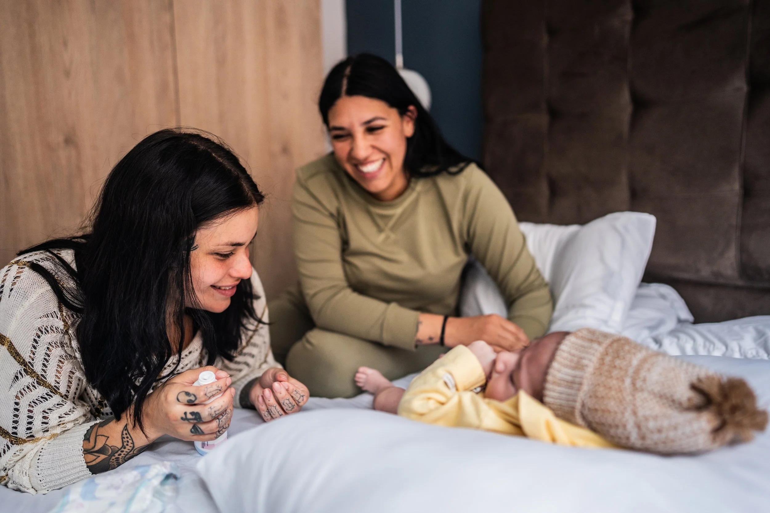 Two women smiling and leaning over a bed to look at a baby lying on the bed. The women appear happy and engaged with the baby.