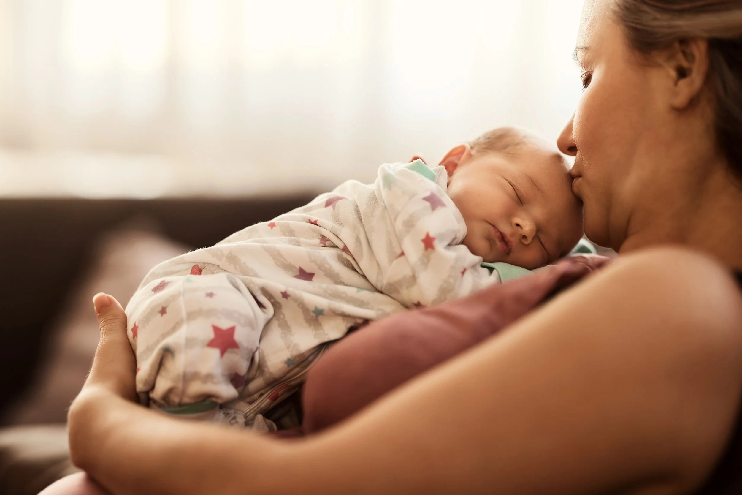 A mother holding her sleeping baby close with foreheads touching in a tender moment indoors.