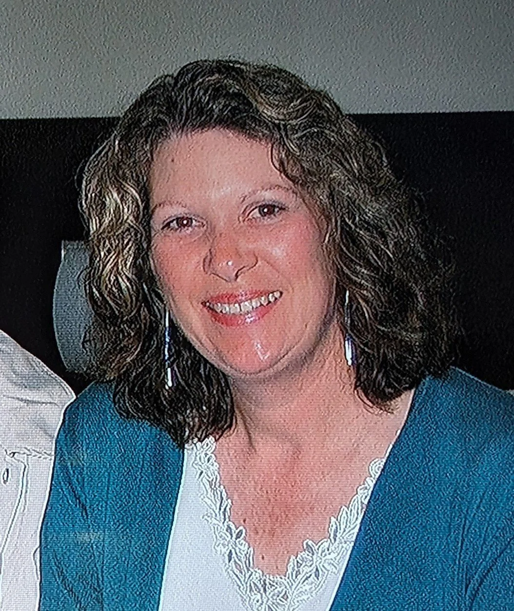 A woman with curly brown hair wearing a teal blazer and white lace top, smiling at the camera.