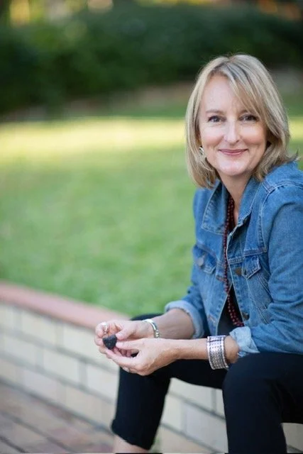 A woman with blonde hair smiling, sitting outdoors on a brick ledge, holding a blackberry in her hands, dressed in a denim jacket over a patterned shirt