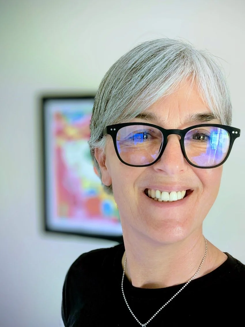 A woman with short gray hair, wearing black glasses and a black top, smiling, with a colorful abstract painting in the background.