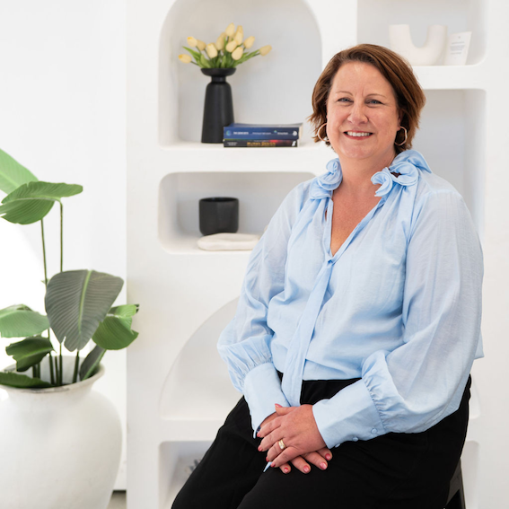 A woman with short brown hair wearing a light blue blouse, sitting and smiling in a modern, white room with shelves, a black vase with yellow flowers, books, a black container, and a large green potted plant.
