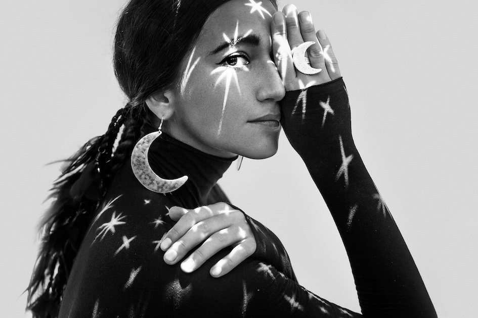 A woman with dark hair styled in braids, wearing large crescent moon earrings and a black turtleneck, poses with her hand covering one eye. Her face and clothing are decorated with star and moon patterns, and star-shaped light reflections are projected on her skin.