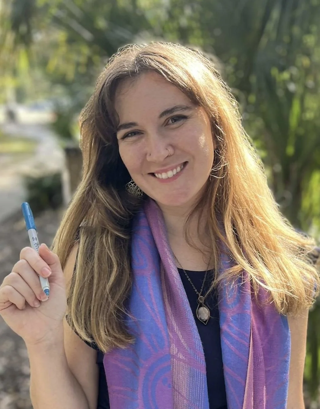 A woman with long, wavy light brown hair and a nose piercing, smiling outdoors, holding a blue pen, wearing a purple scarf and a black top.