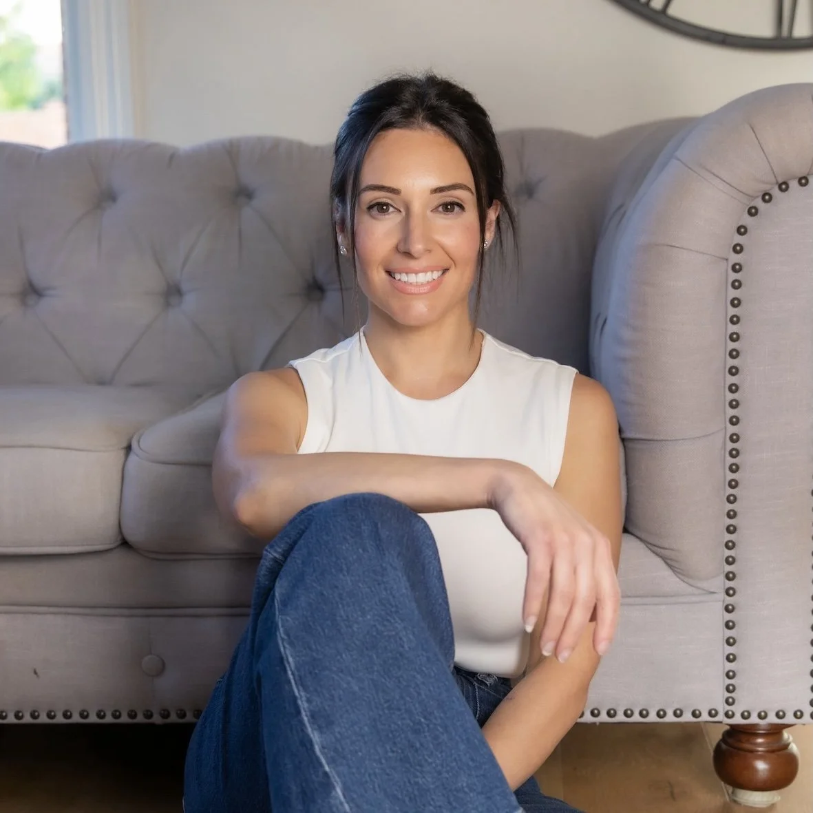 A woman with dark hair sitting on the floor in front of a beige sofa, smiling, wearing a white sleeveless top and blue jeans.