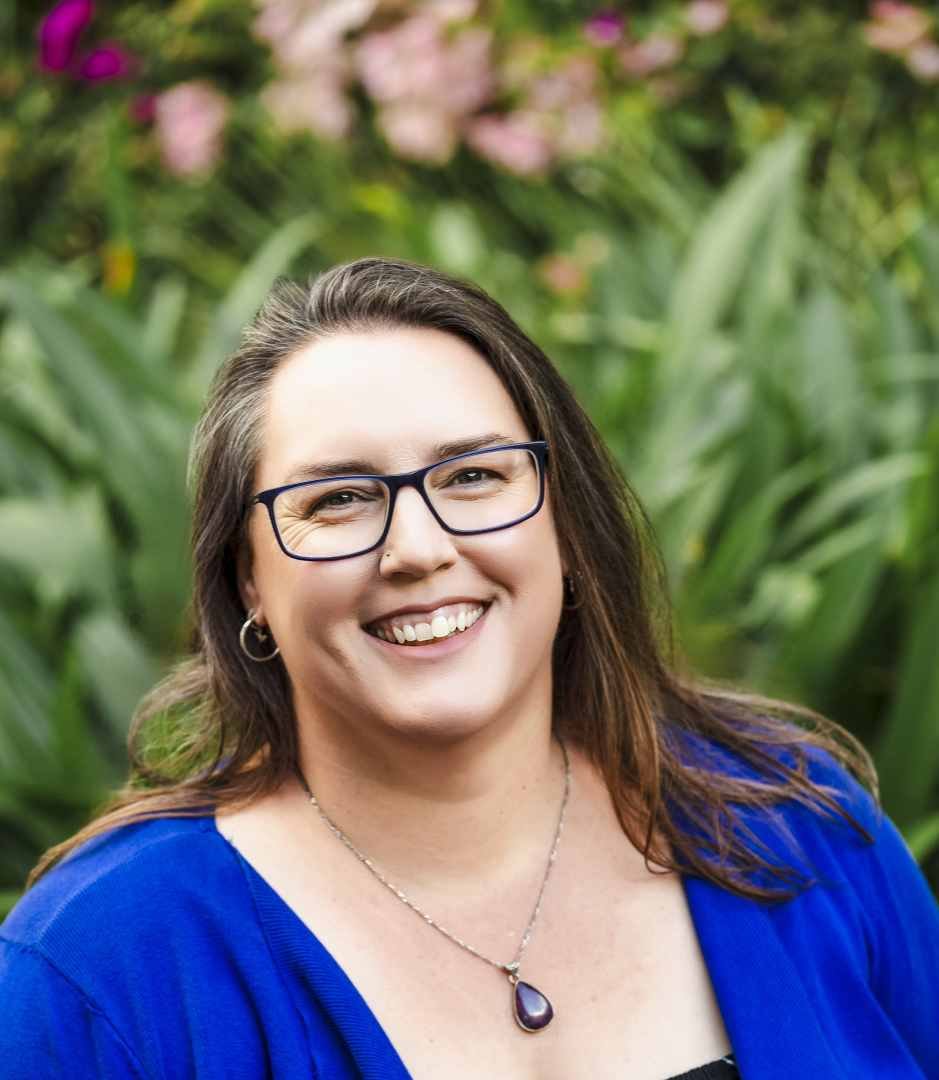 Smiling woman with glasses and earrings, wearing a blue top and a necklace, outdoors in front of green plants and colourful flowers.
