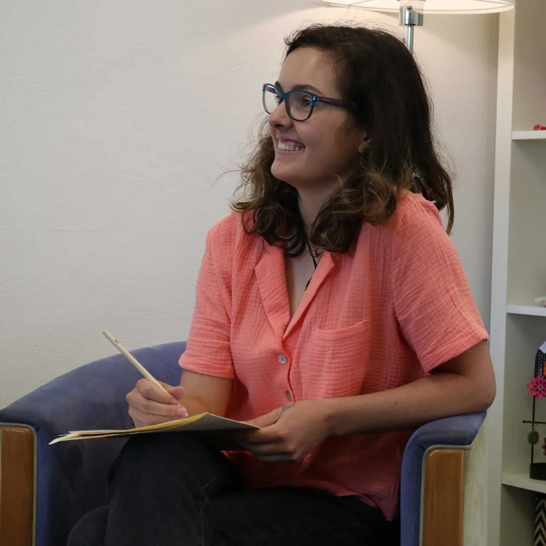 A woman with shoulder-length dark curly hair and glasses, wearing an orange short-sleeve shirt, sitting on a blue chair with wooden armrests, holding a notebook and pencil, smiling and looking to her right, in a room with a shelf and white wall behind her.
