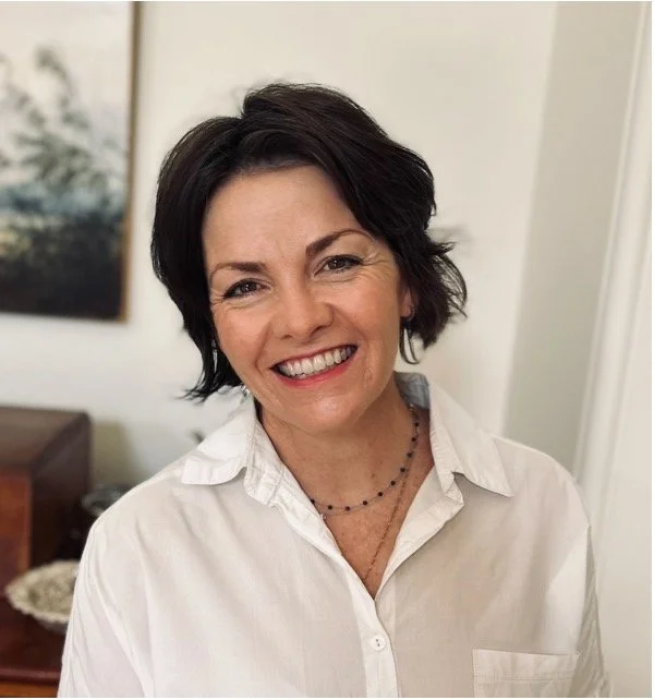 Smiling woman with short dark hair wearing a white blouse and a black beaded necklace, standing indoors.