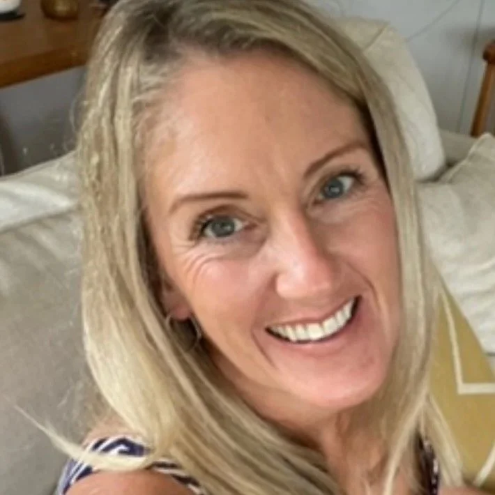 A smiling woman with blonde hair and blue eyes taking a selfie on a bed inside a room.