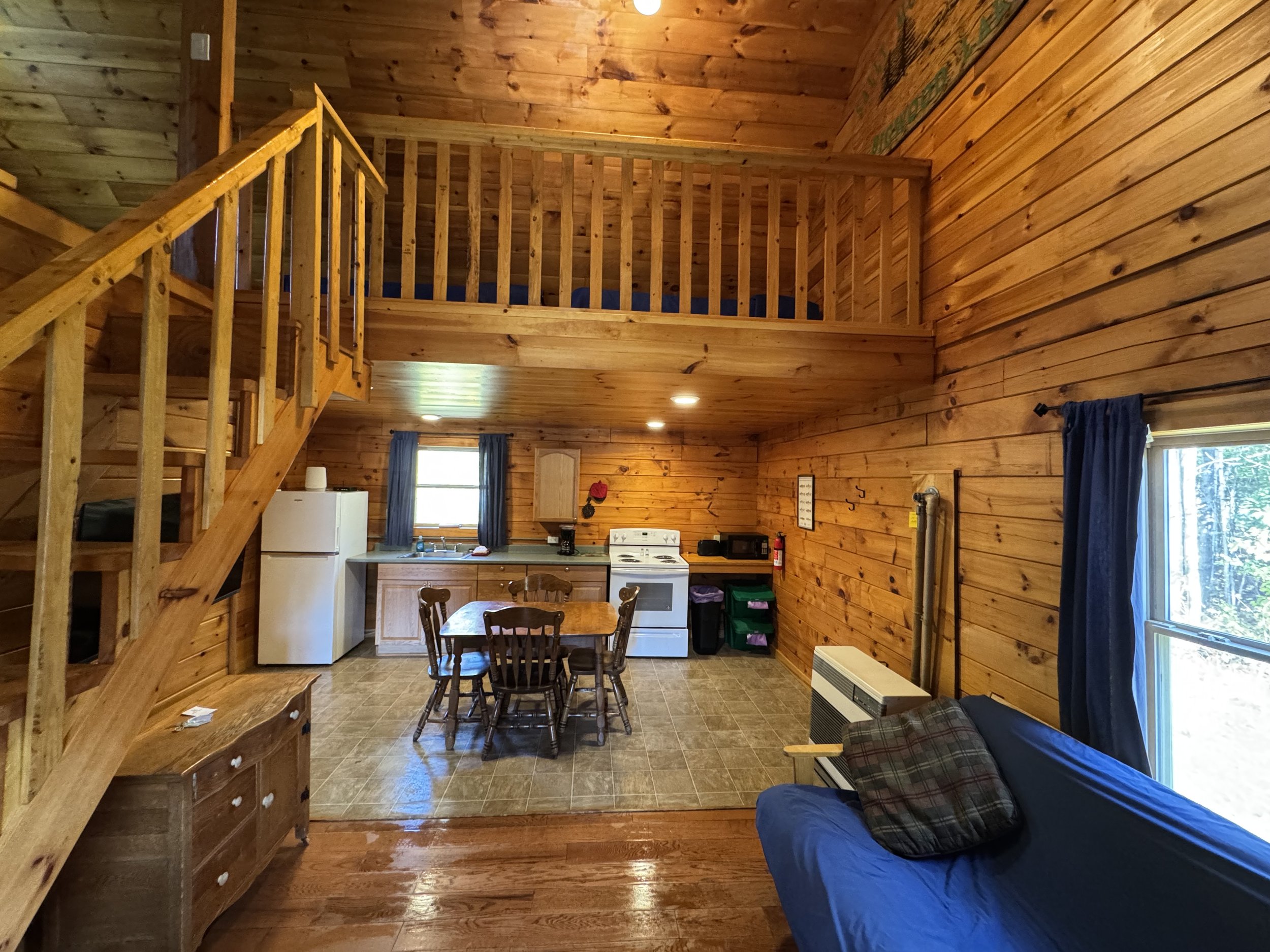 Interior view of a cabin with wood-paneled walls, a staircase leading to an upper loft, a small kitchen area with a dining table, a window with blue curtains, a couch with a pillow, and appliances including a refrigerator, stove, microwave, and an ai