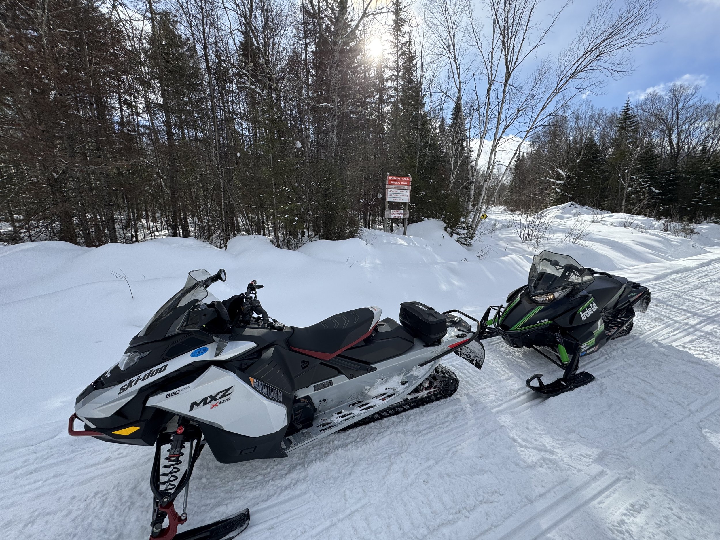 A snow-covered forest with two snowmobiles parked on the trail, a white one on the left and a black and green one on the right, under a partly cloudy sky with the sun shining through the trees.