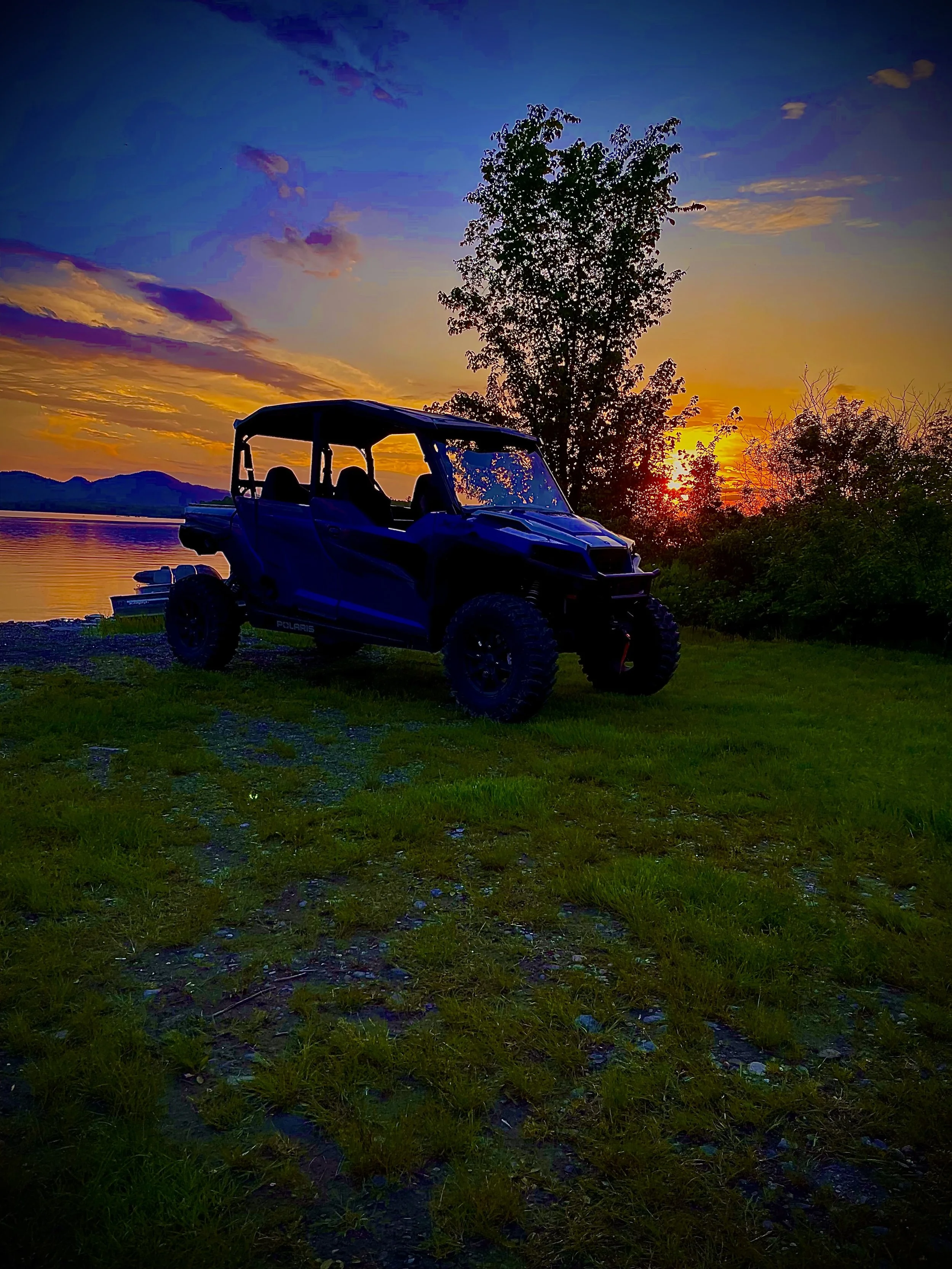 A black off-road side-by-side vehicle parked on grassy ground next to a lake at sunset, with trees and mountains in the background and a colorful sky.
