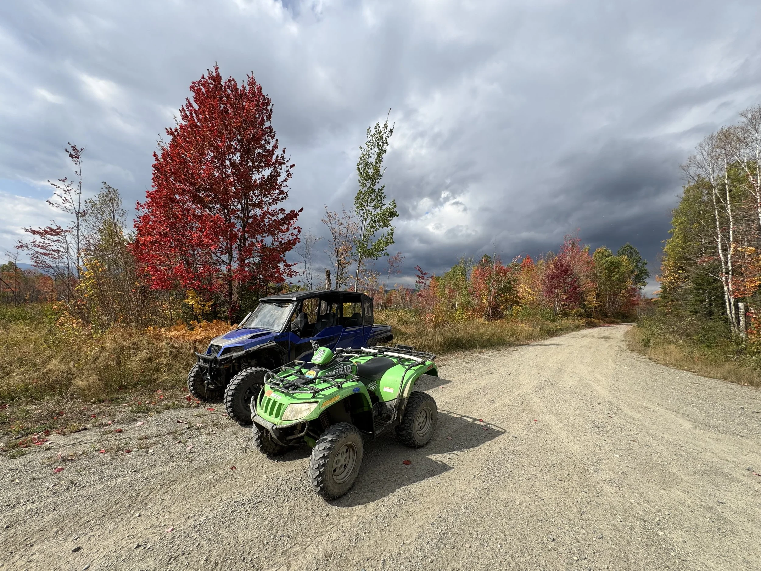 Two all-terrain vehicles (ATVs), one green and one blue, parked on a gravel dirt trail near the Border Riders Clubhouse in a forested area with colorful autumn trees and a cloudy sky.