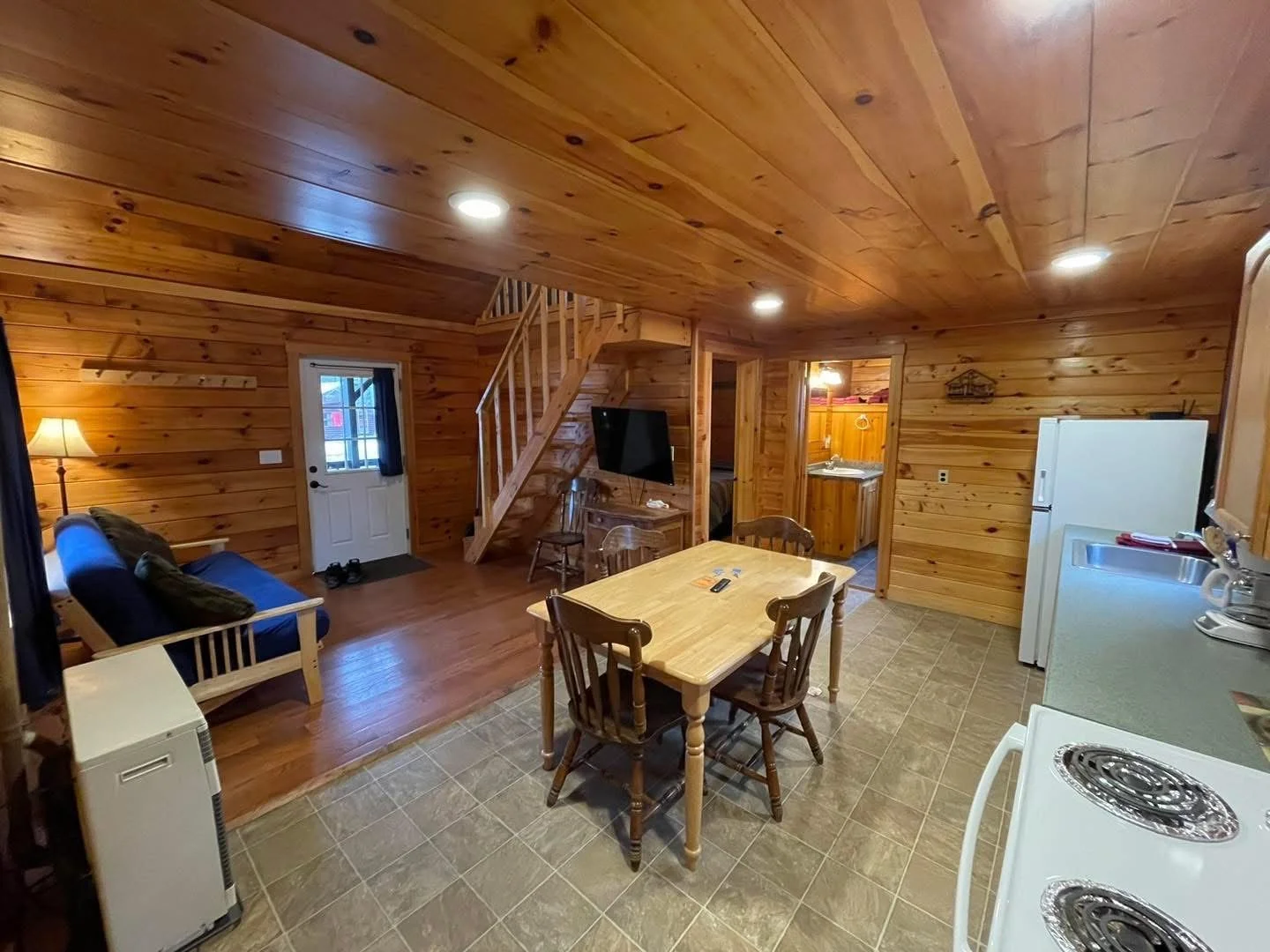 Interior view of a cozy, rustic cabin kitchen and living area with wooden walls and ceiling, a dining table with chairs, a sofa, a TV, a refrigerator, a stove, and a door leading outside.