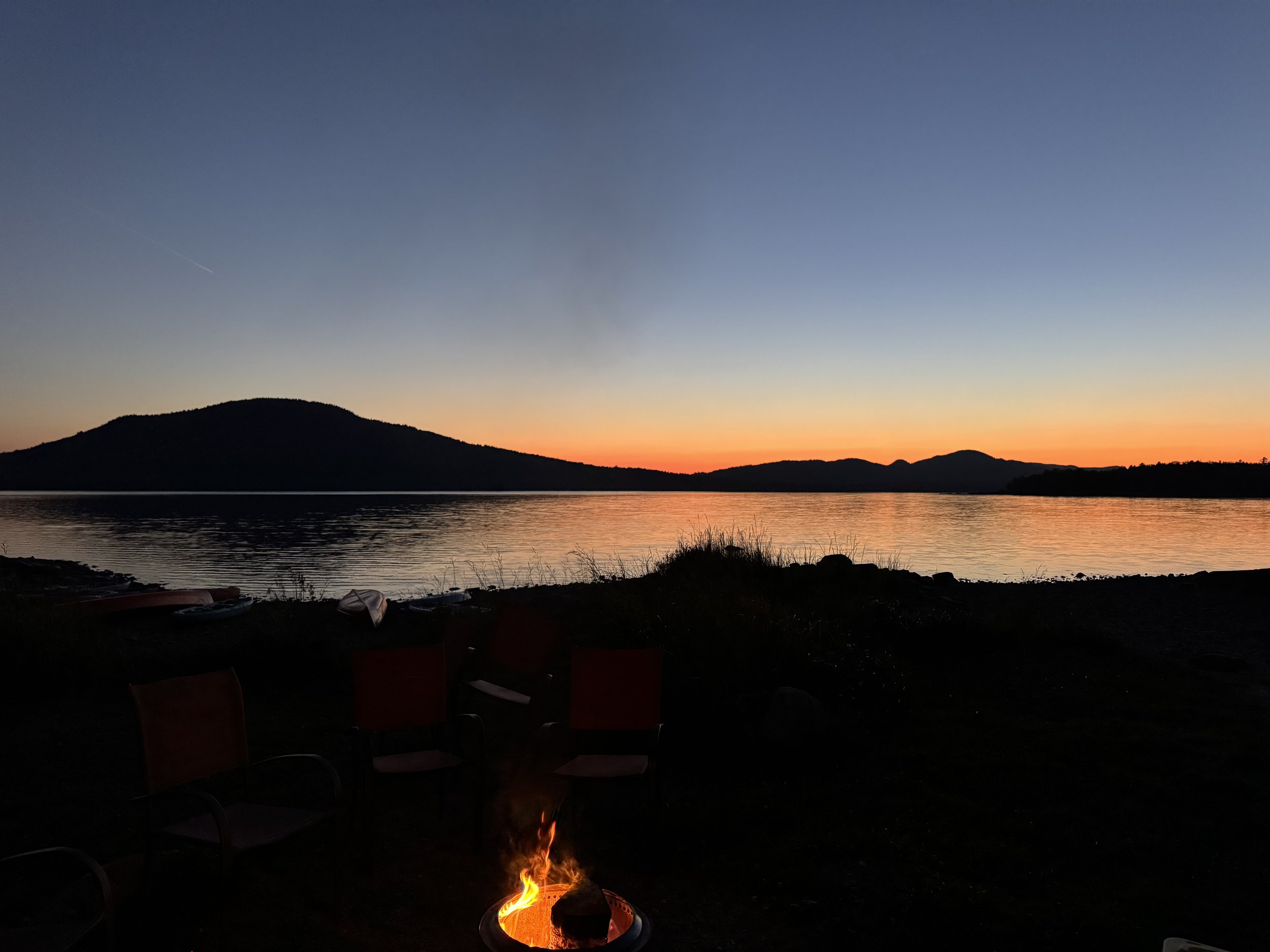 Sunset over Big Wood lake with a silhouette of Sally Mountain and Attean in the background, and chairs with a small campfire in the foreground.