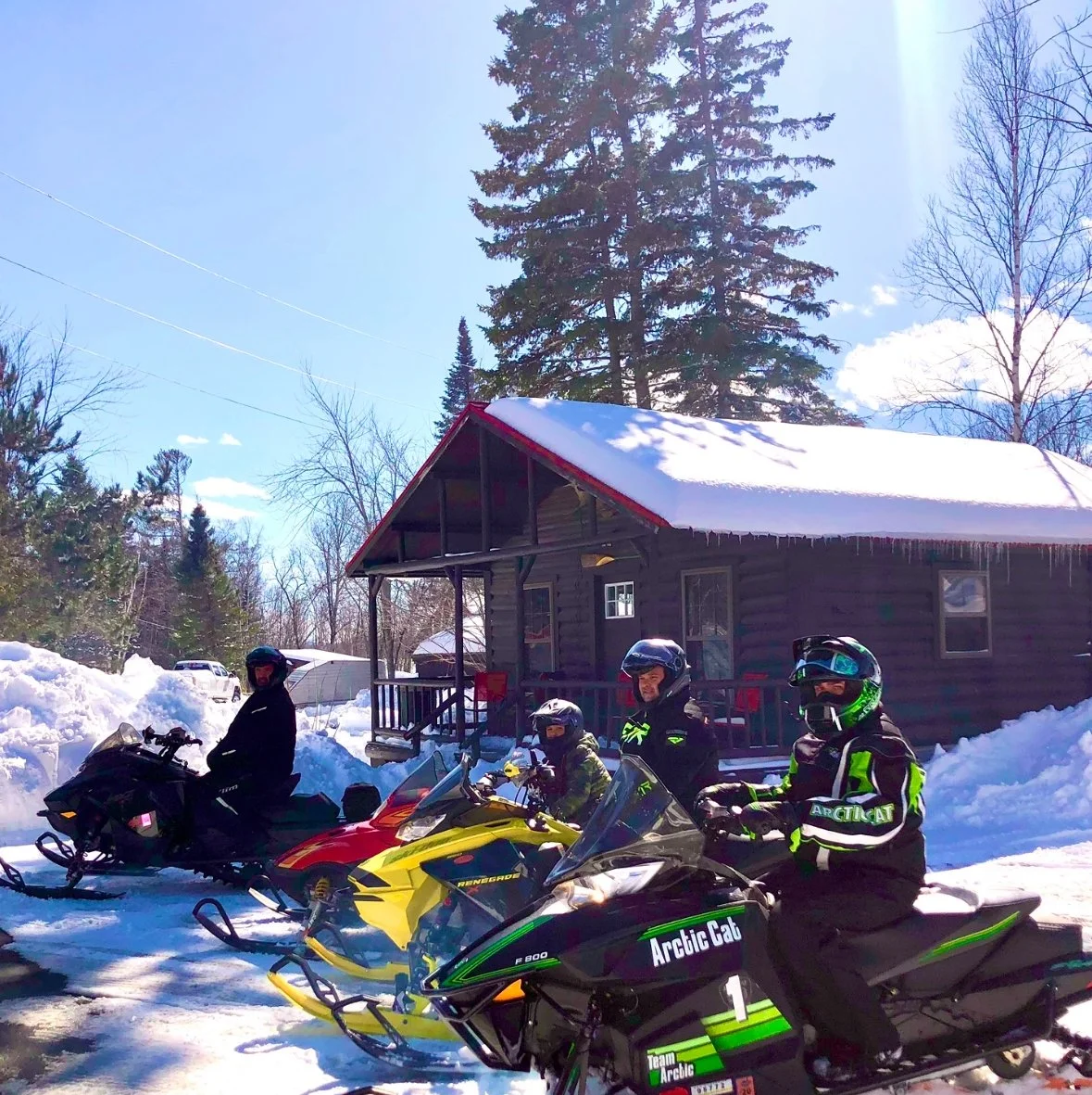 Four people dressed in winter gear sitting on colorful snowmobiles parked in front of Sunrise cabin, a Maine log cabin with snow-covered roof and icicles, on a bright winter day.