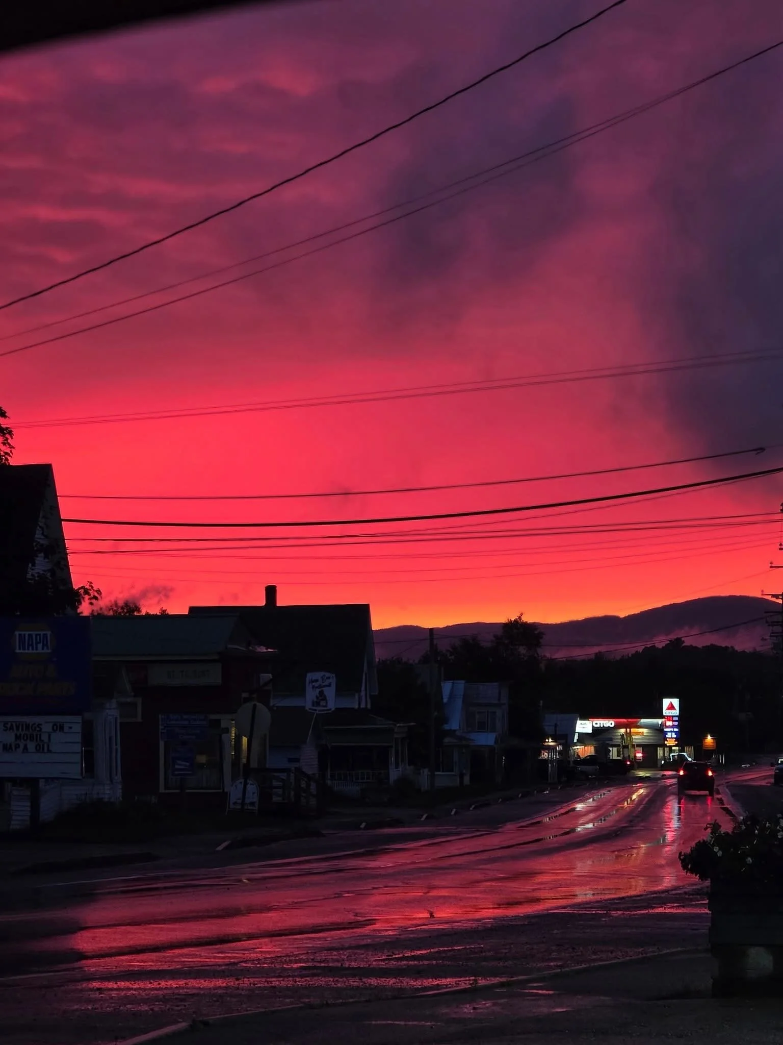 A vibrant sunset sky with shades of pink and purple, reflecting on a wet street Route 201 in the small town of Jackman (The Switzerland of Maine) with houses and Bishops Store gas station in the distance.