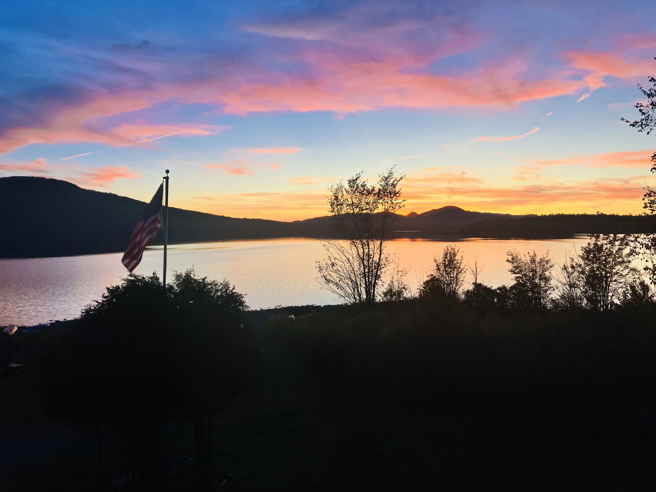 Sunset over a lake with orange, pink, and purple clouds, silhouette trees and an American flag on a flagpole in the foreground.