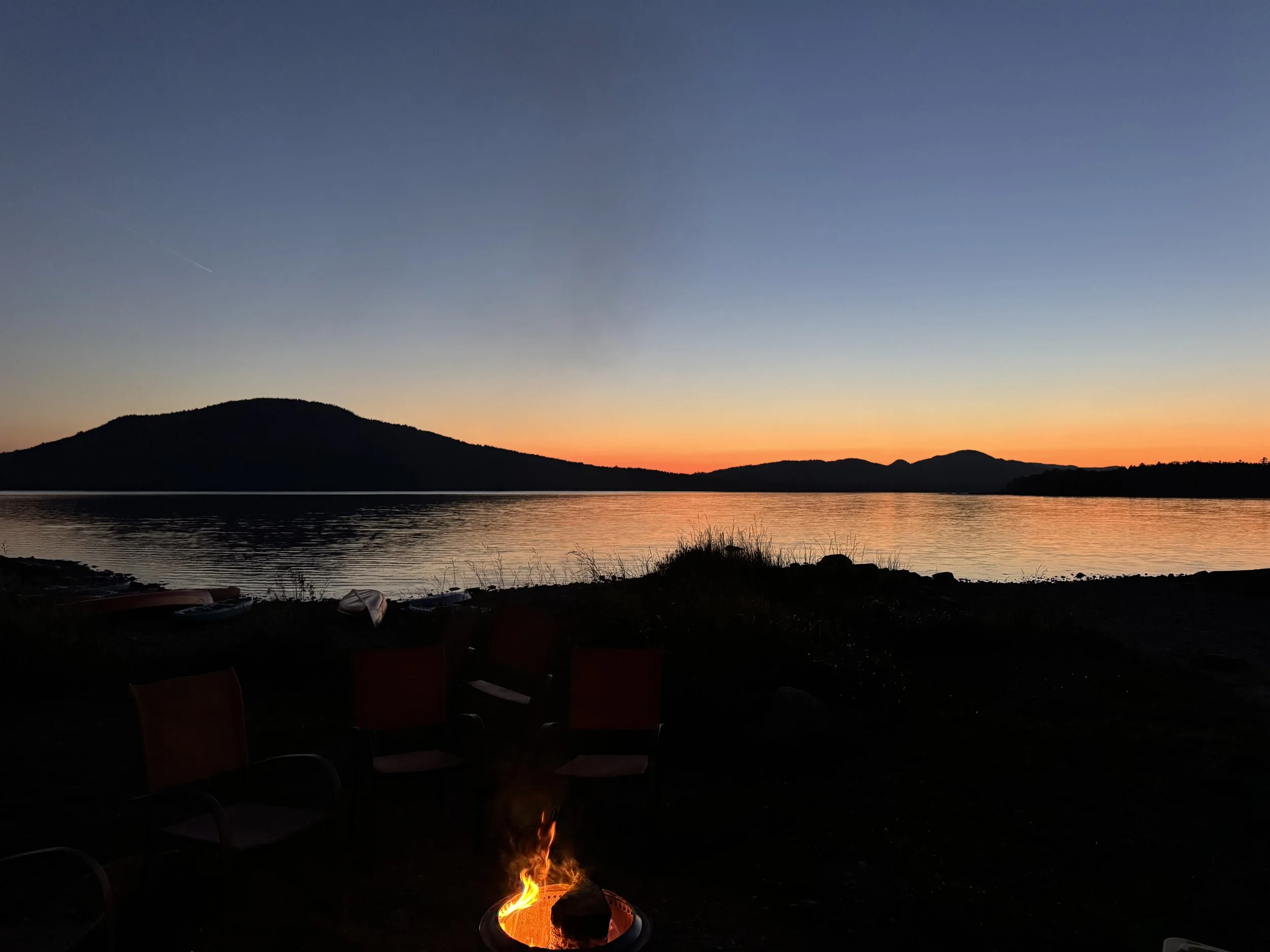 A sunset over a lake with distant hills, with a small campfire and chairs in the foreground.