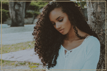 A young woman with long curly dark hair looking down, standing outdoors near trees and grass.
