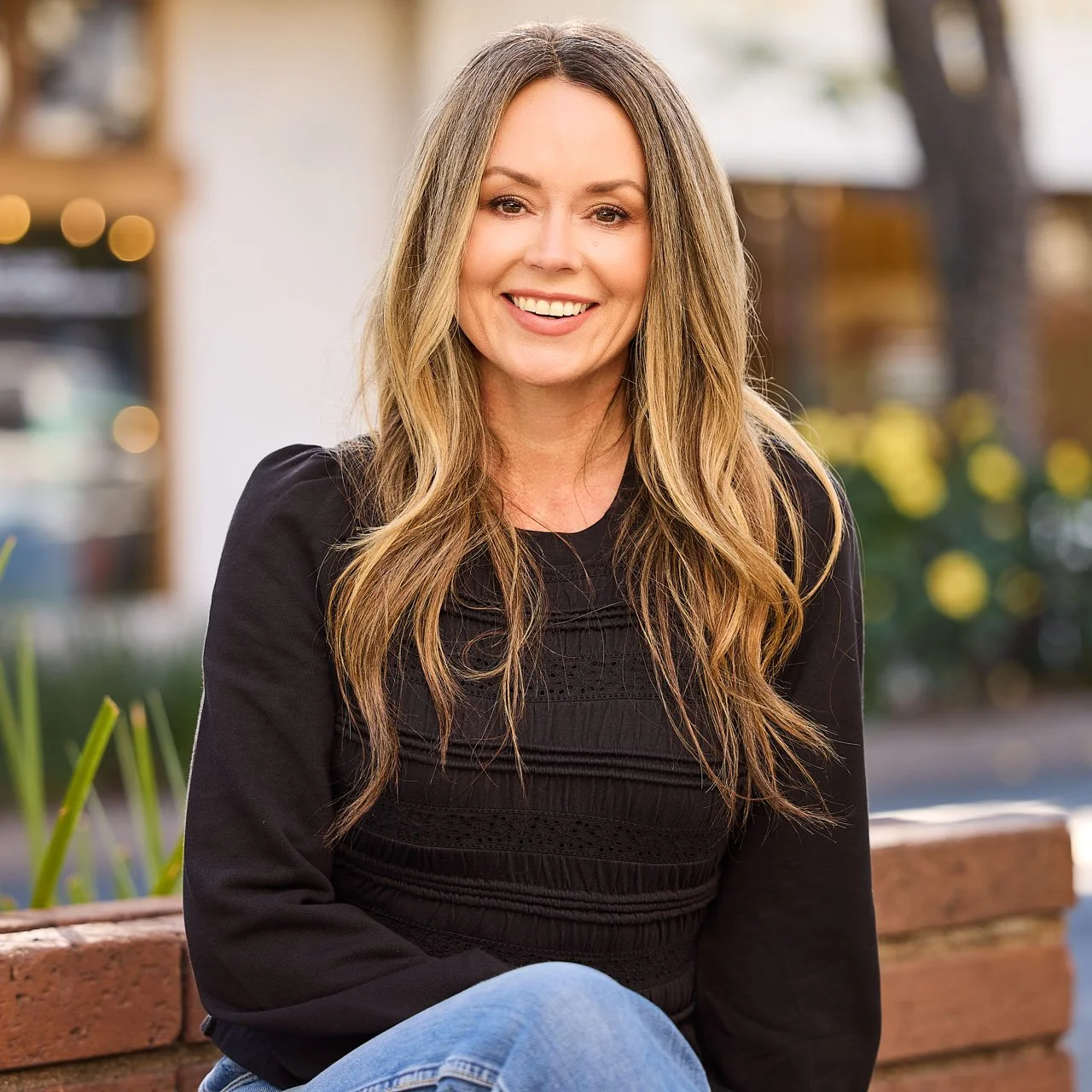 A woman with long, wavy blonde hair, smiling, sitting on a brick bench outdoors with blurred background of flowers and trees.
