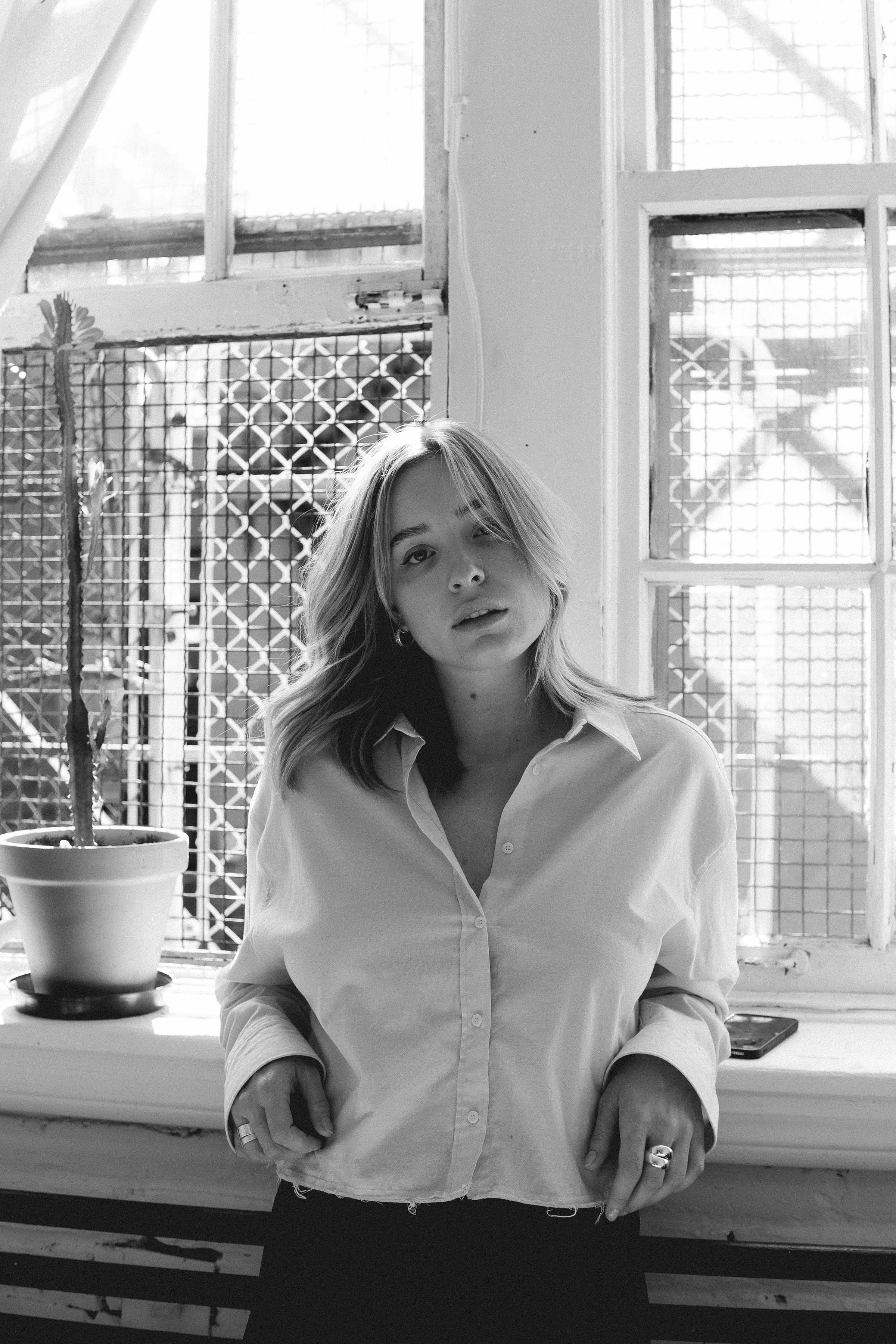 Black and white photo of a woman with shoulder-length hair, wearing a slightly oversized button-up shirt, standing in front of a window with sunlight behind her, and a potted cactus plant on the windowsill.