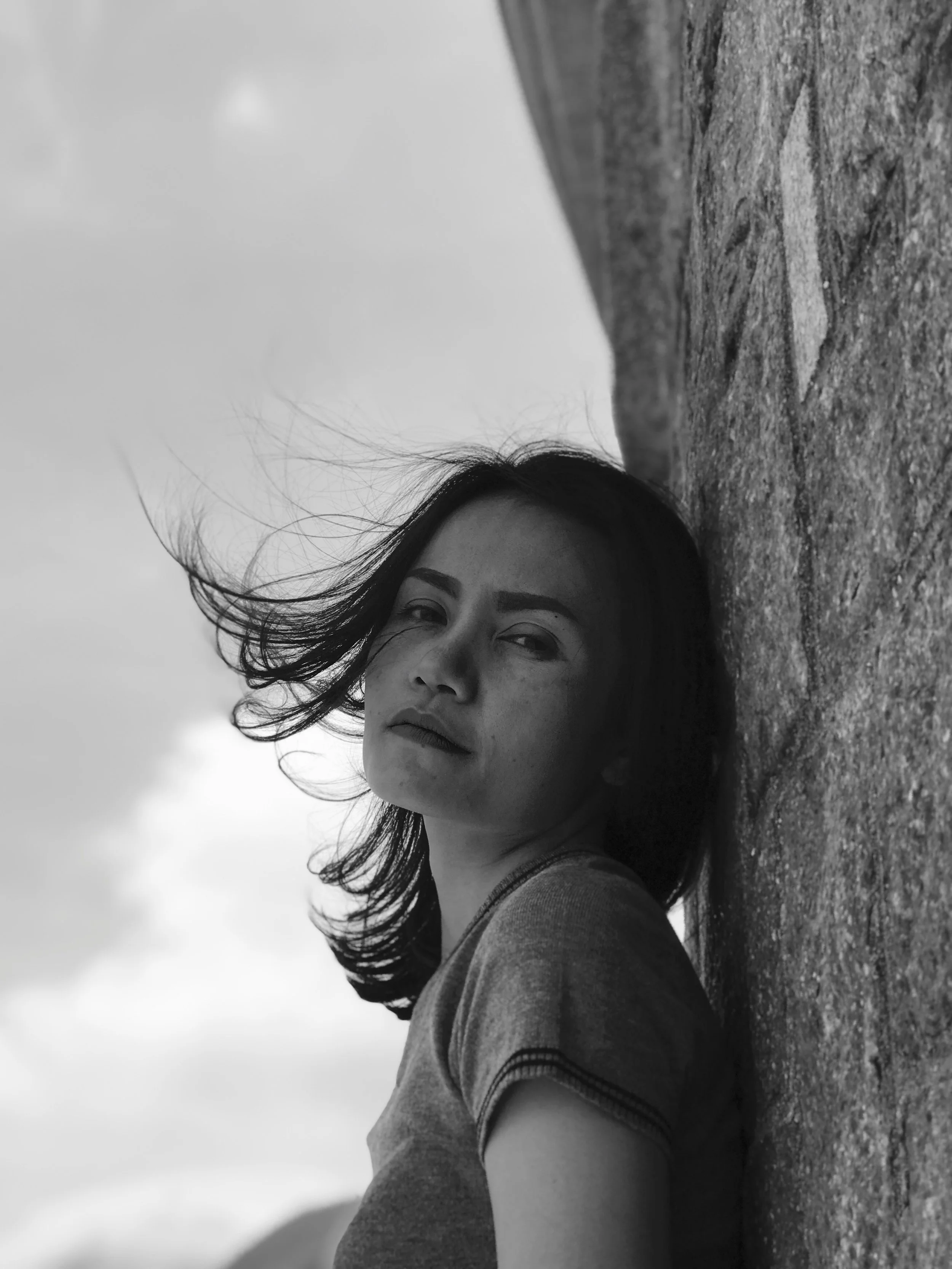 A black-and-white photo of a woman with dark hair outdoors, leaning against a stone wall with her head tilted back. Her hair is blown by the wind, and she has a calm, serious expression.