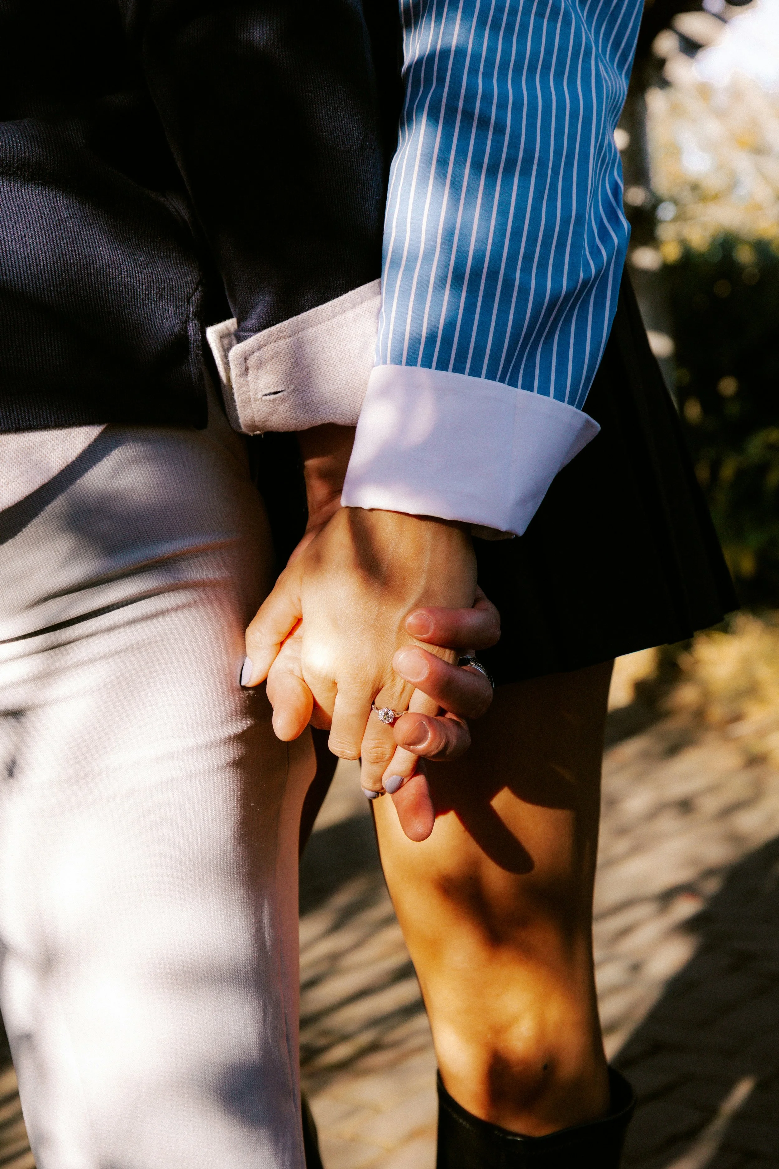 Two people holding hands, with one wearing a ring, outdoors with sunlight and blurred natural background.