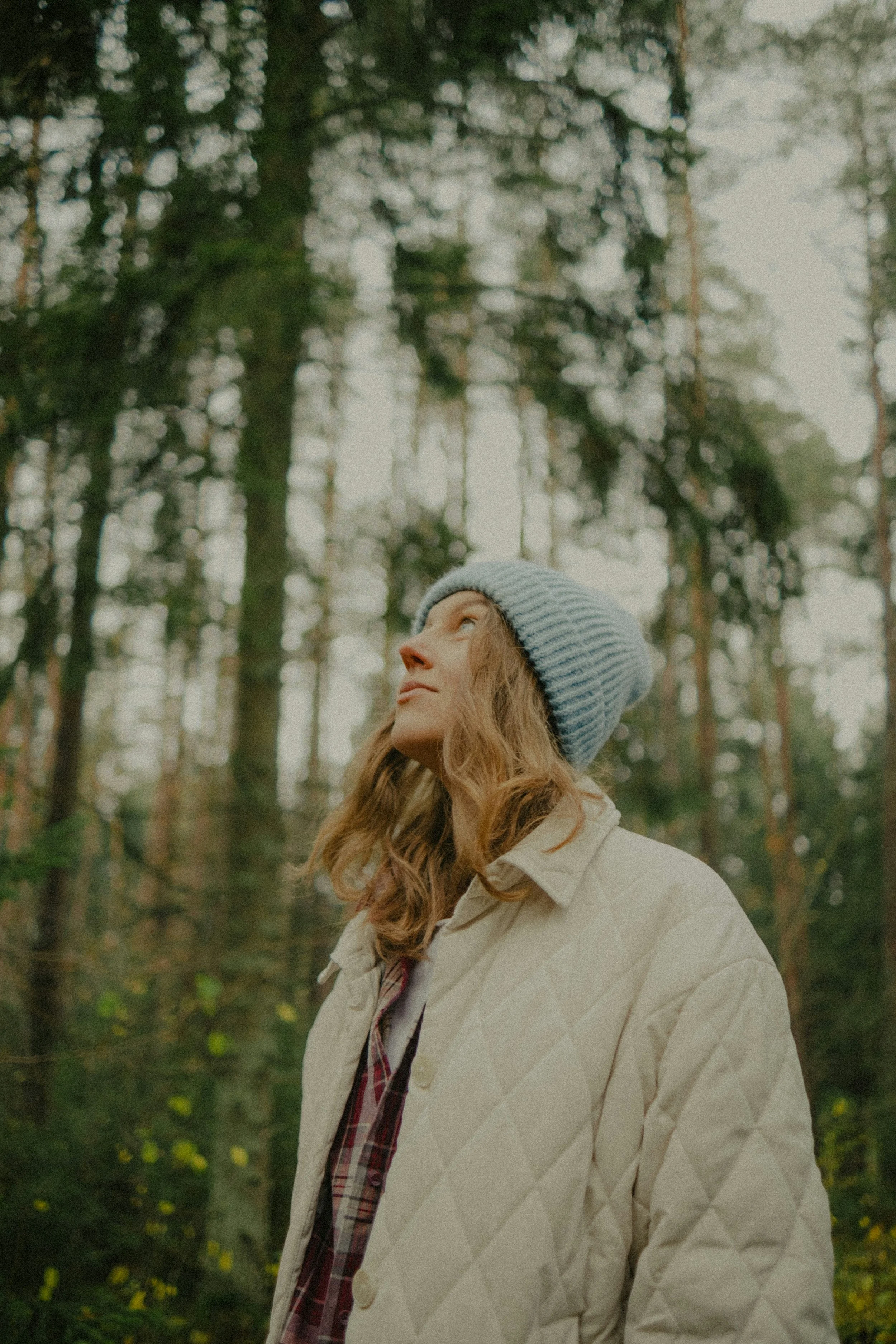 A woman with wavy brown hair wearing a blue knit beanie, a beige quilted jacket, and a plaid shirt, looking upward in a forest setting with tall trees and green foliage.