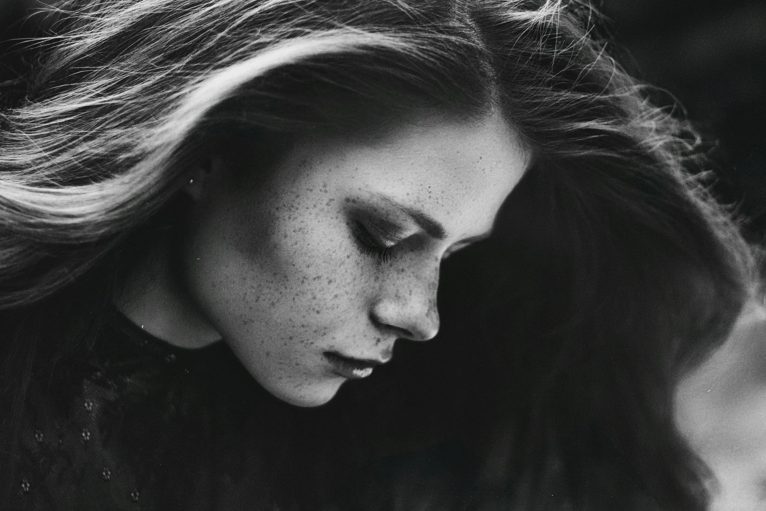 A black and white close-up portrait of a woman with long hair, freckles, and eyes closed, looking downward.