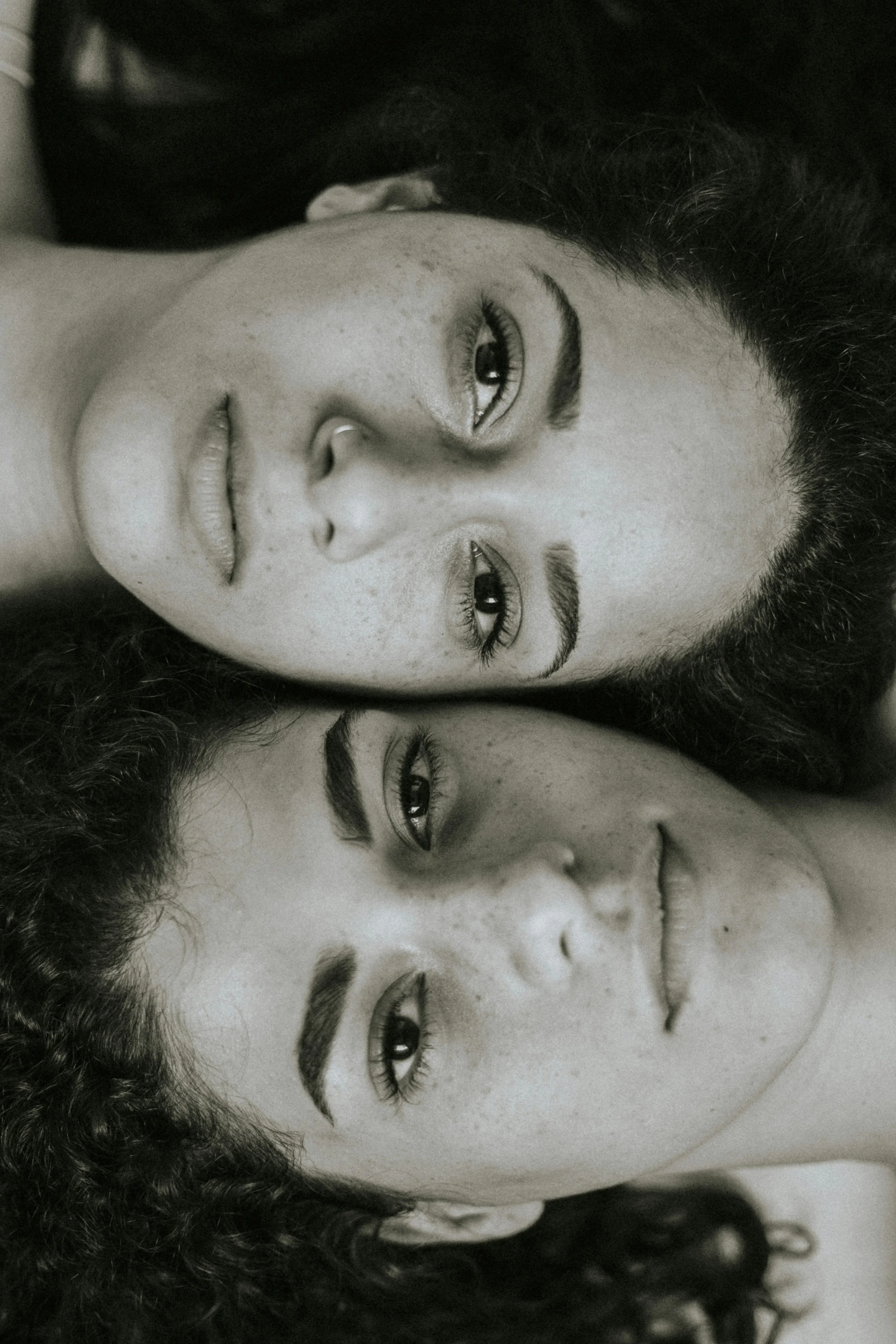 Close-up black and white photo of two women lying down, facing the camera, with their heads side by side and slightly tilted, both with curly hair and prominent eyebrows.
