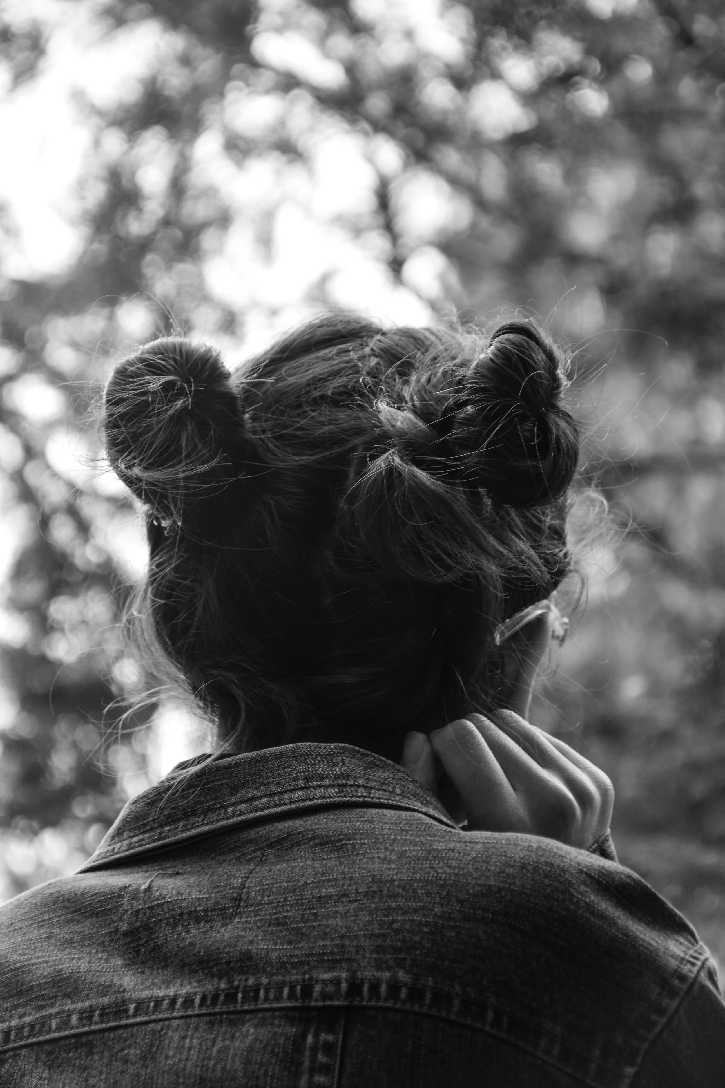 A woman with her back turned, sporting two messy buns and wearing a denim jacket, gazes into a blurred outdoor background with trees.