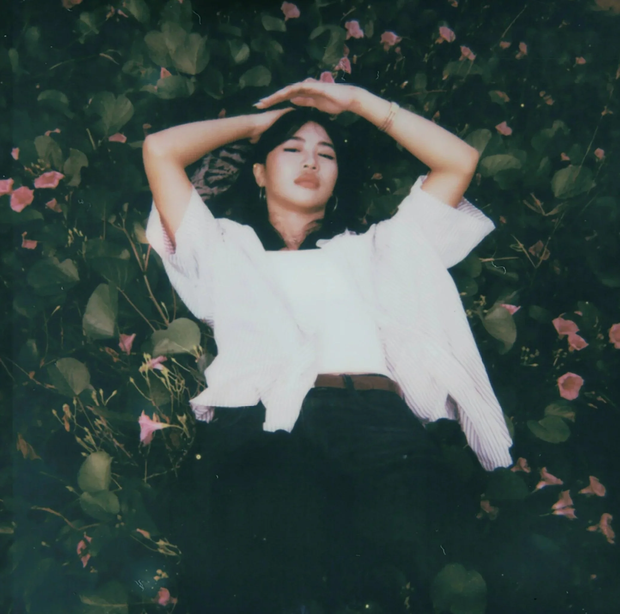 A woman with dark hair and earrings lying on a bed of green leaves and pink flowers, with her arms raised above her head and eyes closed.