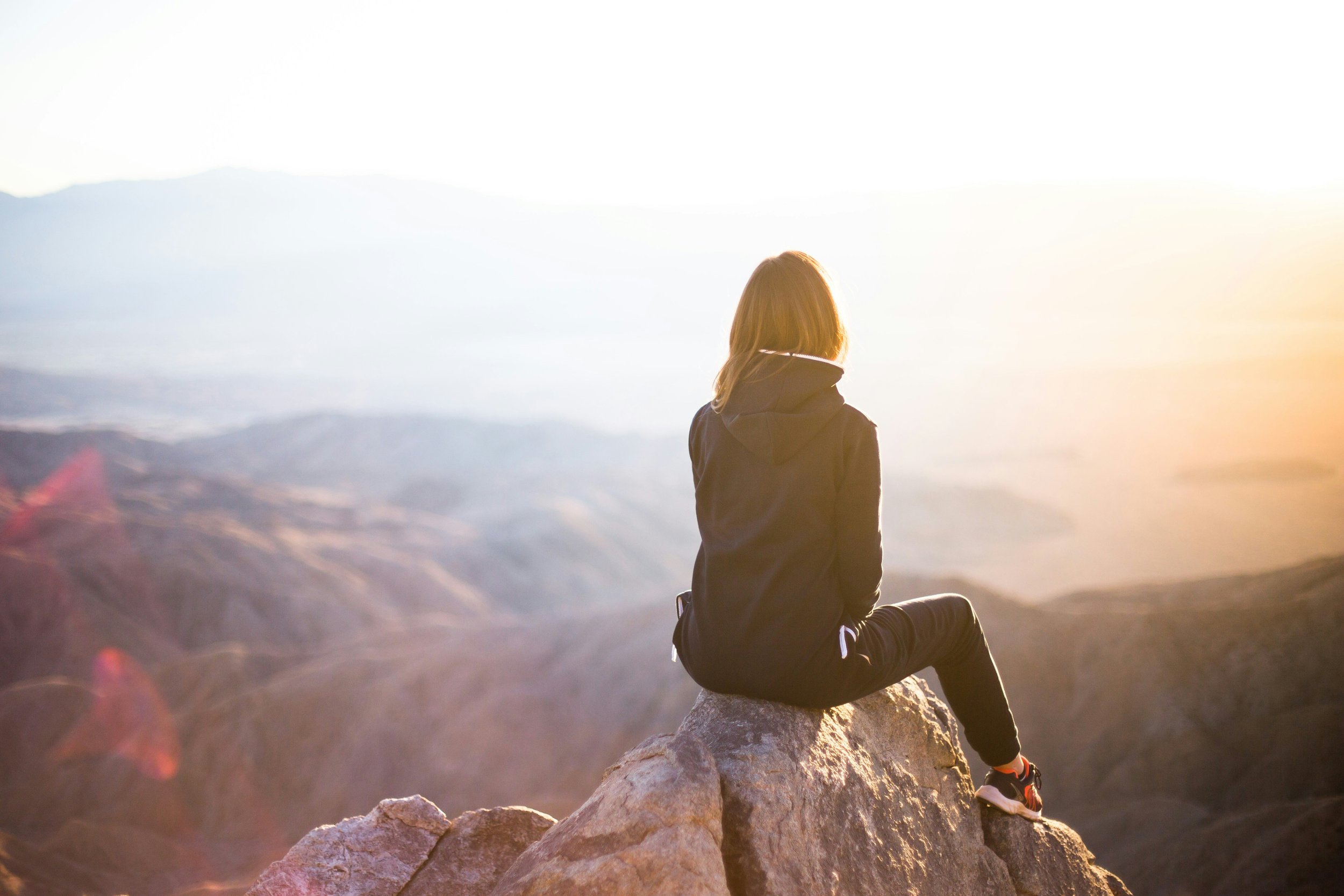 A woman sitting on a rock, facing a mountainscape at sunrise or sunset.