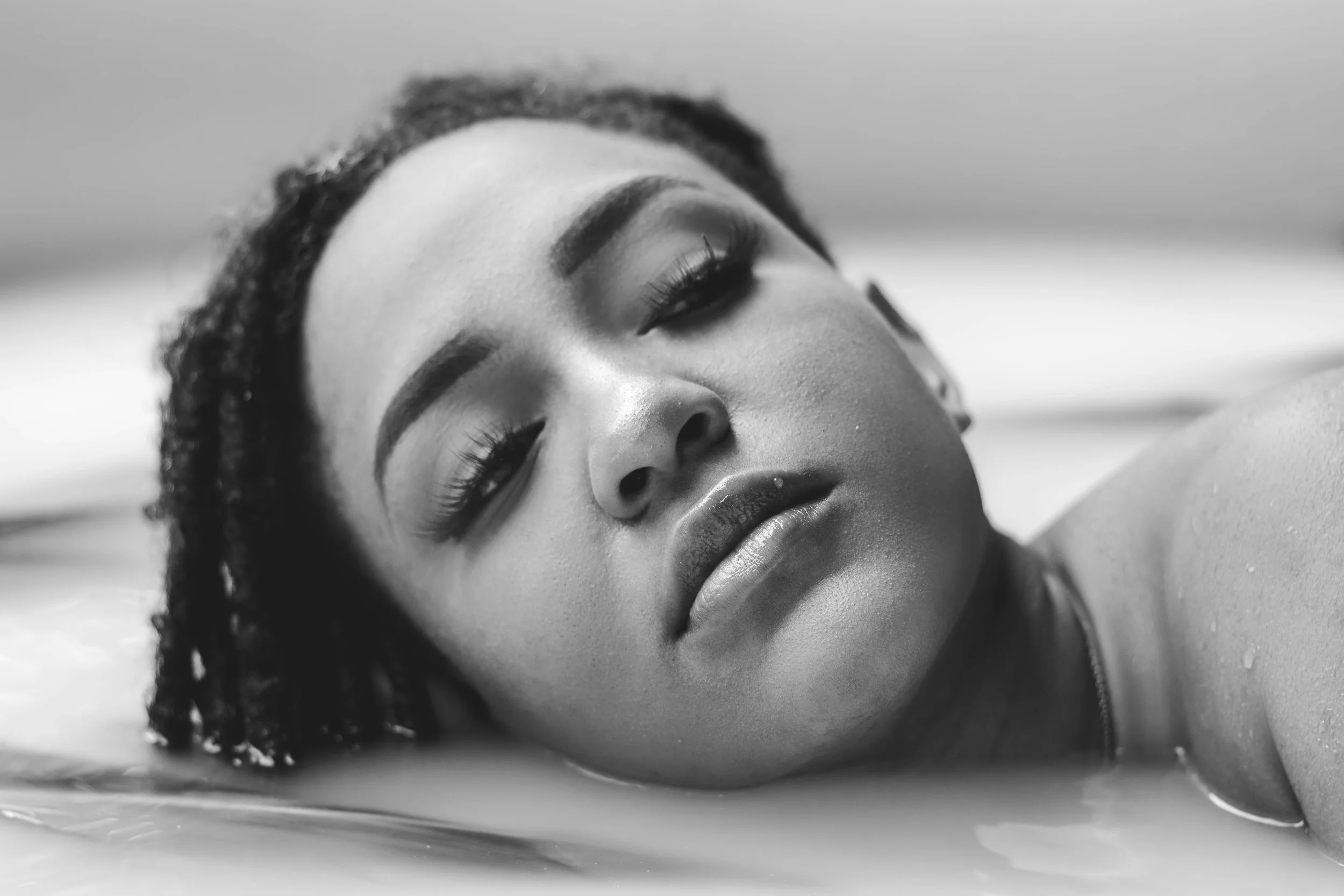 A black-and-white close-up of a woman with closed eyes, lying on her side, wet hair, and relaxed facial expression.