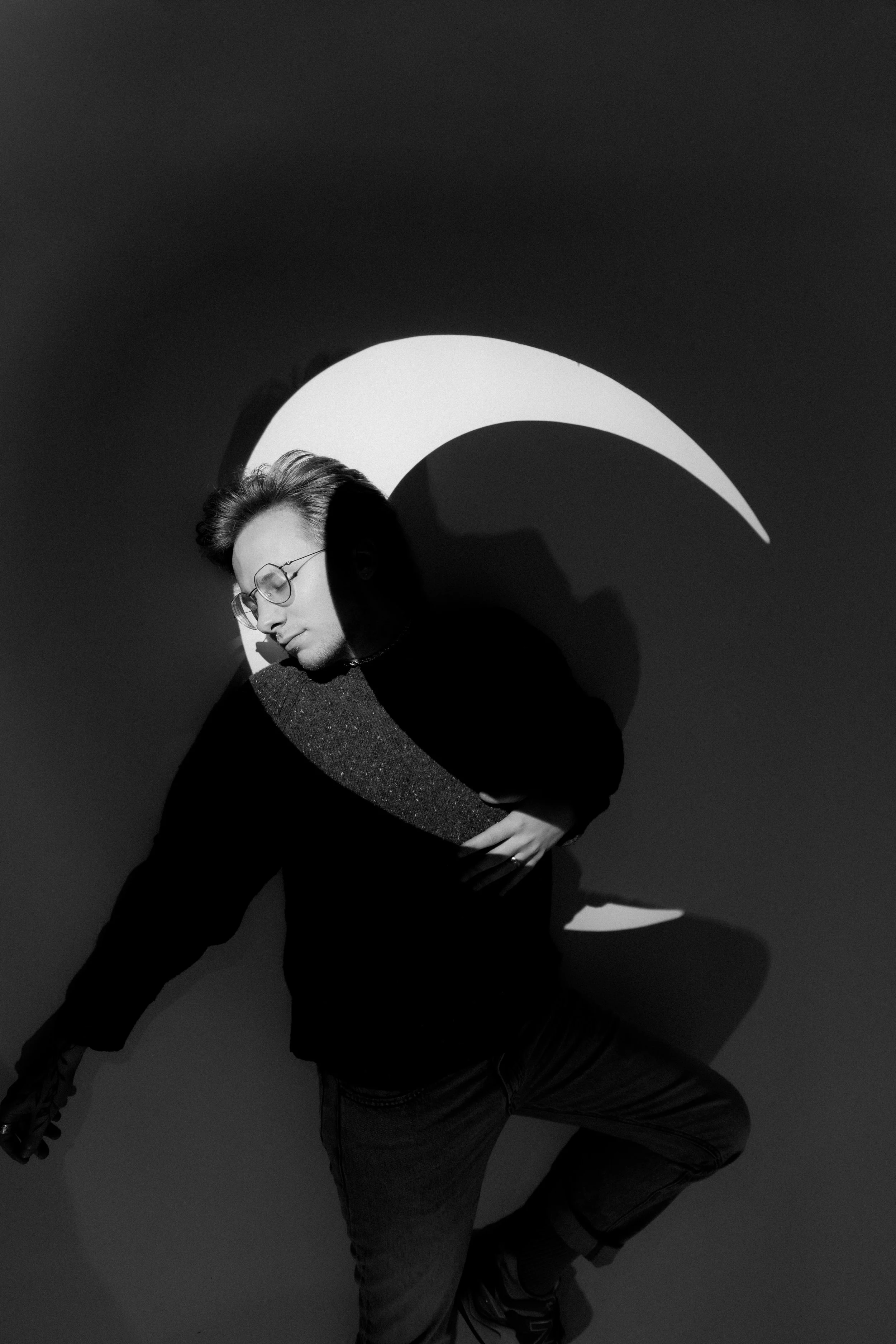 A black-and-white photo of a person with glasses, wearing a hoodie, standing in front of a wall with a moon-shaped light projection. The person is posing with one leg bent and hand on their stomach, with their face turned downward and eyes closed.
