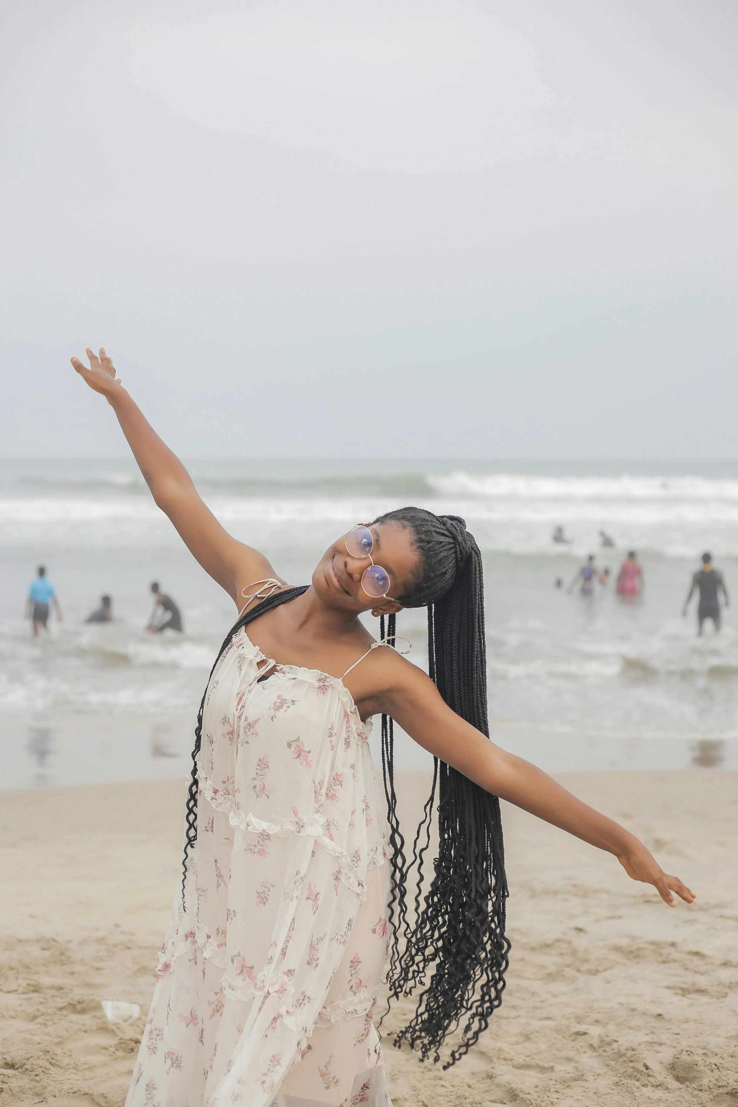 A woman with long braids, wearing glasses and a floral dress, standing on a beach with her arms outstretched in a dance pose, with people swimming and playing in the ocean in the background.