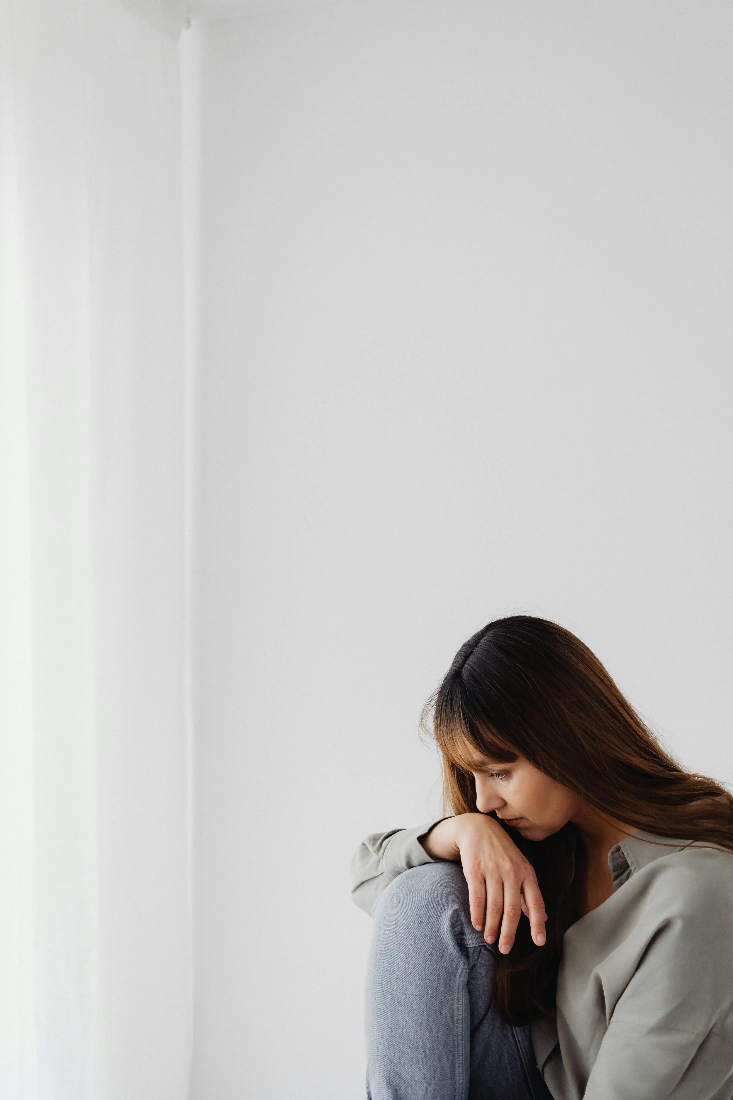 A woman with brown hair sitting on the floor with her head resting on her knee, looking down thoughtfully, against a plain white wall.