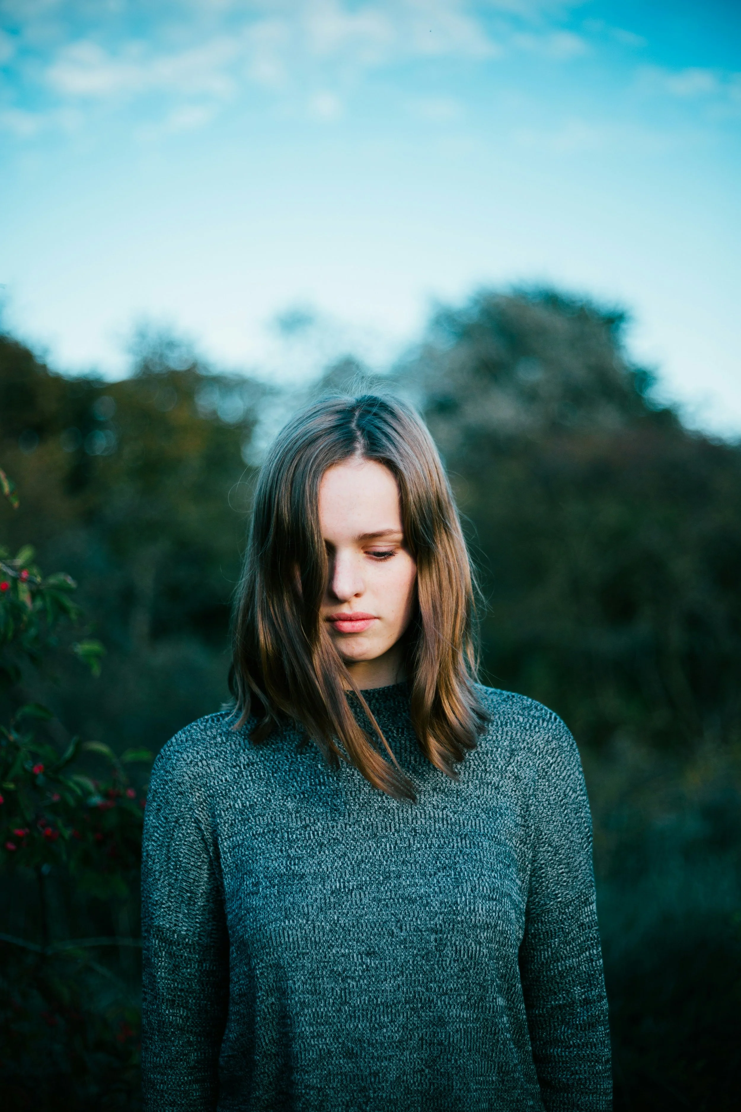 A young woman with shoulder-length brown hair looking downward outdoors during daytime, with trees and a blue sky in the background.