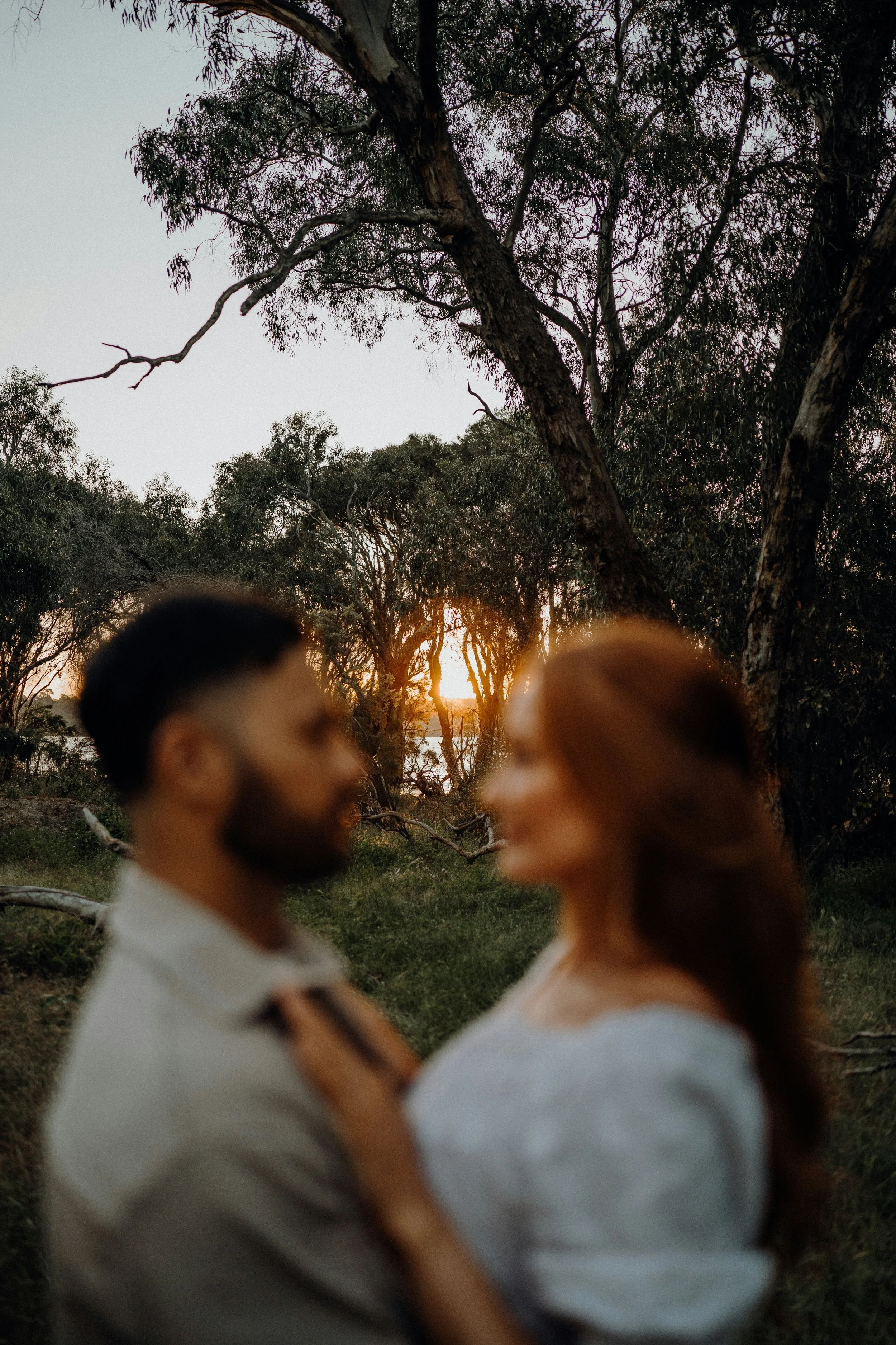 A blurry man and a blurry woman standing close together outdoors with trees and a sunset in the background.
