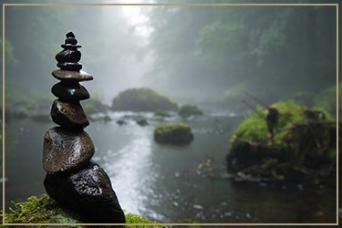 Stacked rocks near a river with moss-covered rocks and foggy forest in the background.