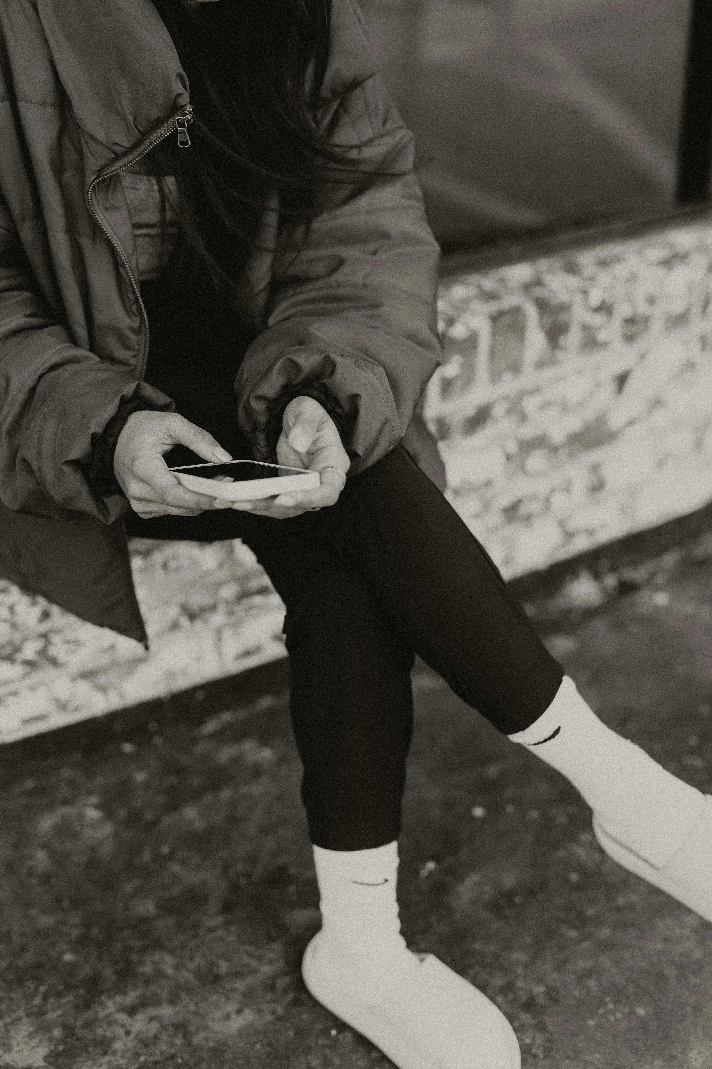 Person sitting outdoors, wearing a jacket, black pants, white Nike socks, and white sneakers, holding a smartphone.